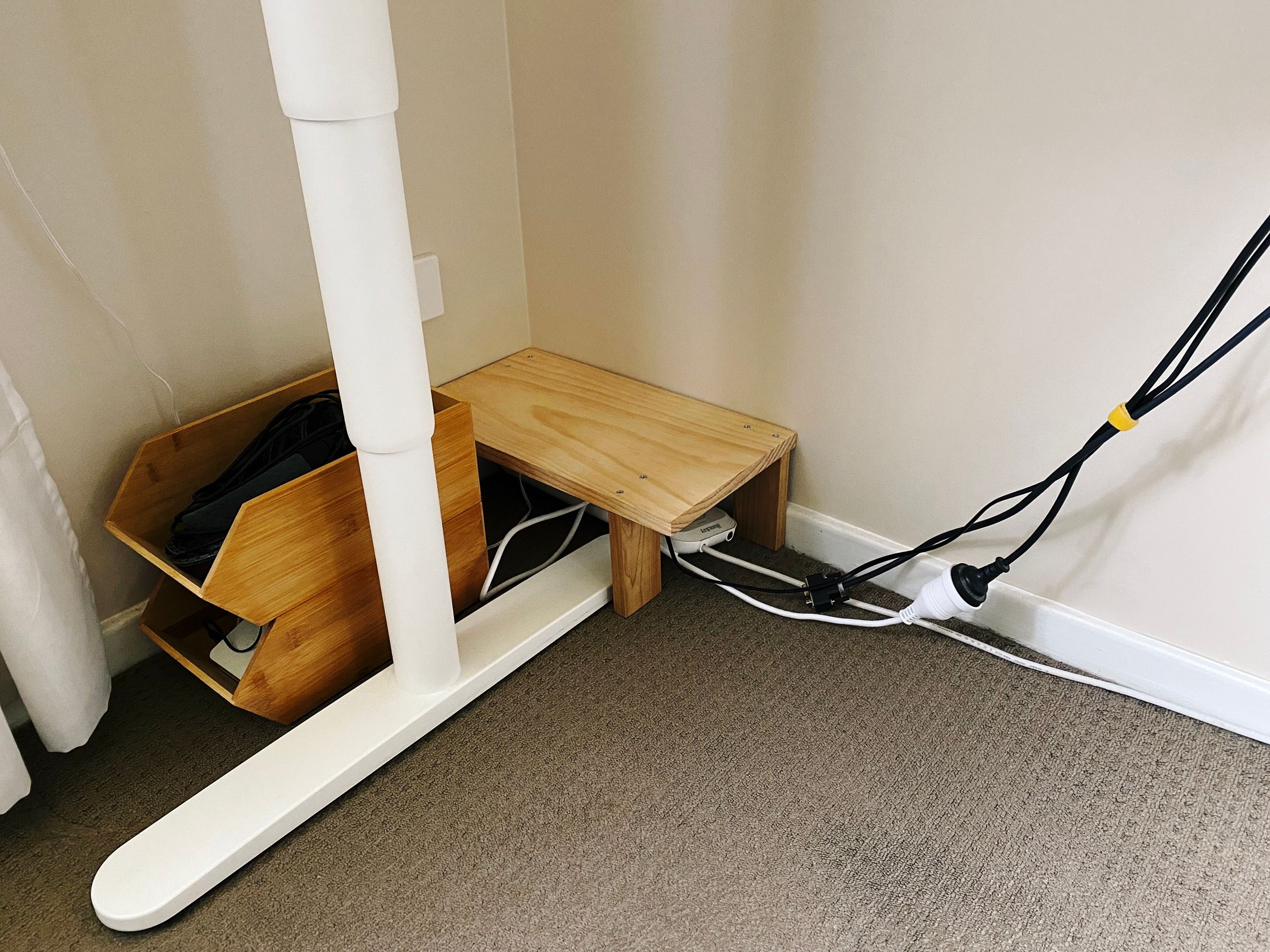 A photo of the corner of a room underneath a standing desk, with a little pine wooden platform on four legs that sits over the top of a power board. The platform itself is about 40cm wide and 23cm deep, and is raised up 13cm off the floor.