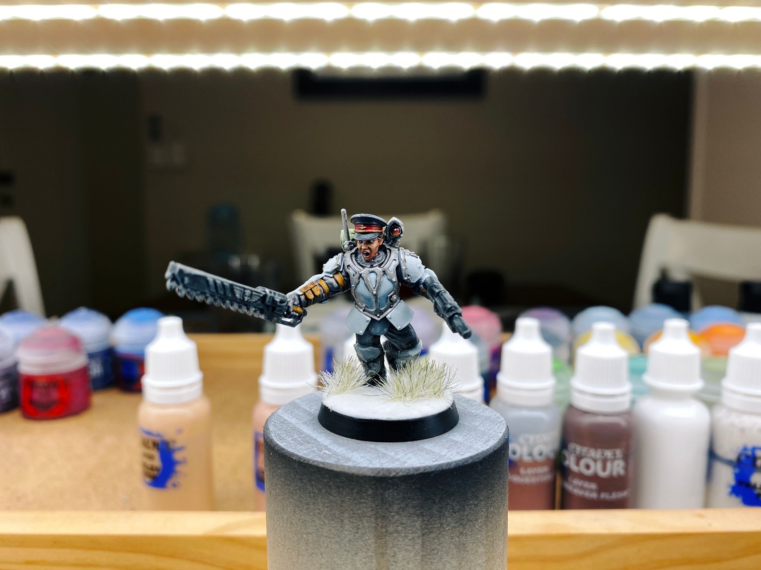 A photo of an Astra Militarum Scion from Warhammer 40,000, basically a regular human trooper except I've replaced the regular male head with a third-party female one that has a military cap on. She's got her mouth open like she's yelling, and is wielding a chainsword and laspistol. The base is all done and looks like snow and it's got a couple of little winter shrubs on it as well.