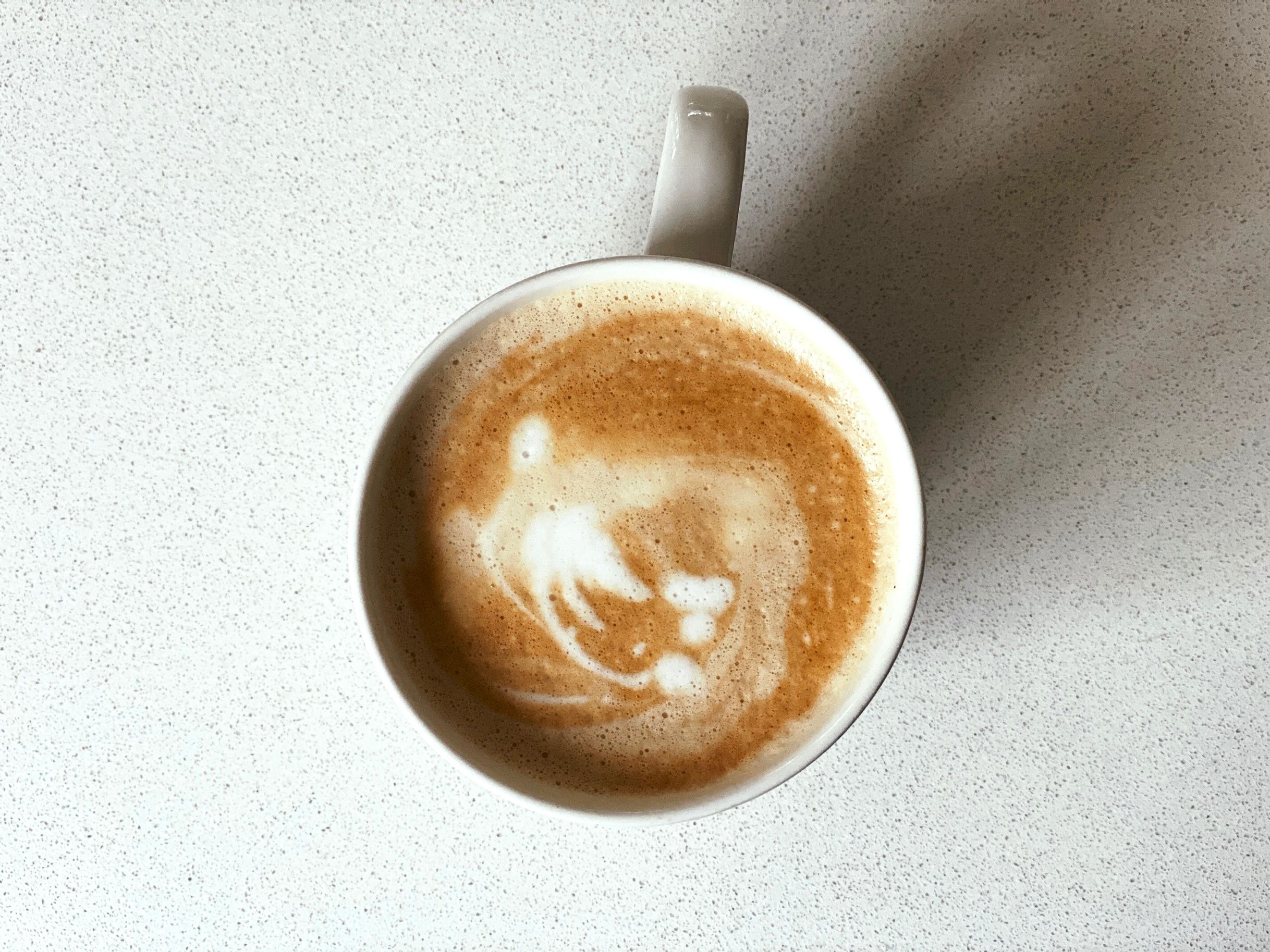 A photo looking down at a the top of a mug of coffee, the foam in the top has what looks like a stylised ghost that looks like it's in motion.