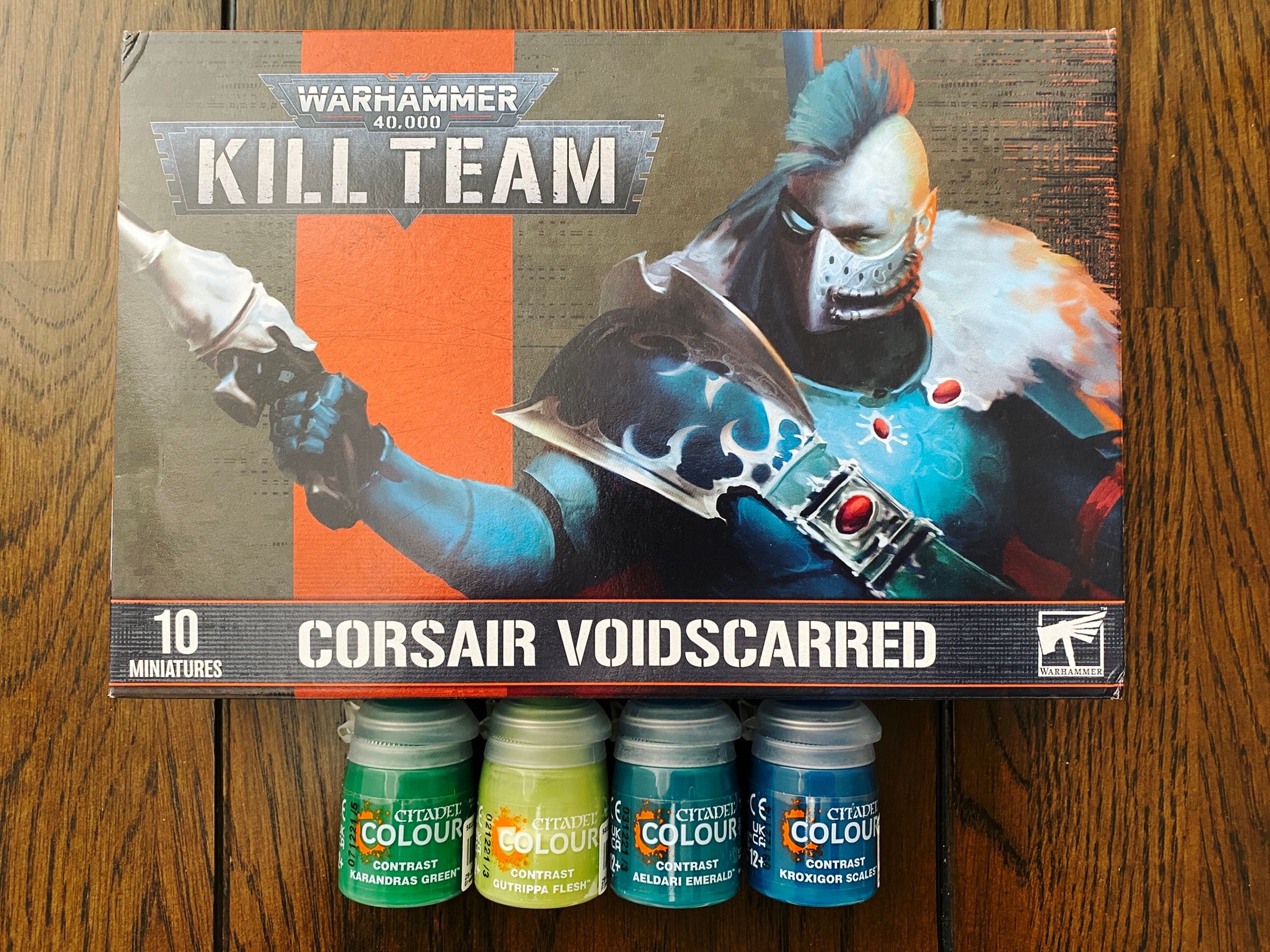 A photo of the Warhammer 40,000 Kill Team squad "Corsair Voidscarred", which are basically space elf pirates, and four pots of paint. The art on the front of the box is of one of the Voidscarred who has a turquoise mohawk and a breathing mask on, and is wearing elegant-looking turquoise armour. He's wielding sleek and elegant-looking bone-coloured pistol.