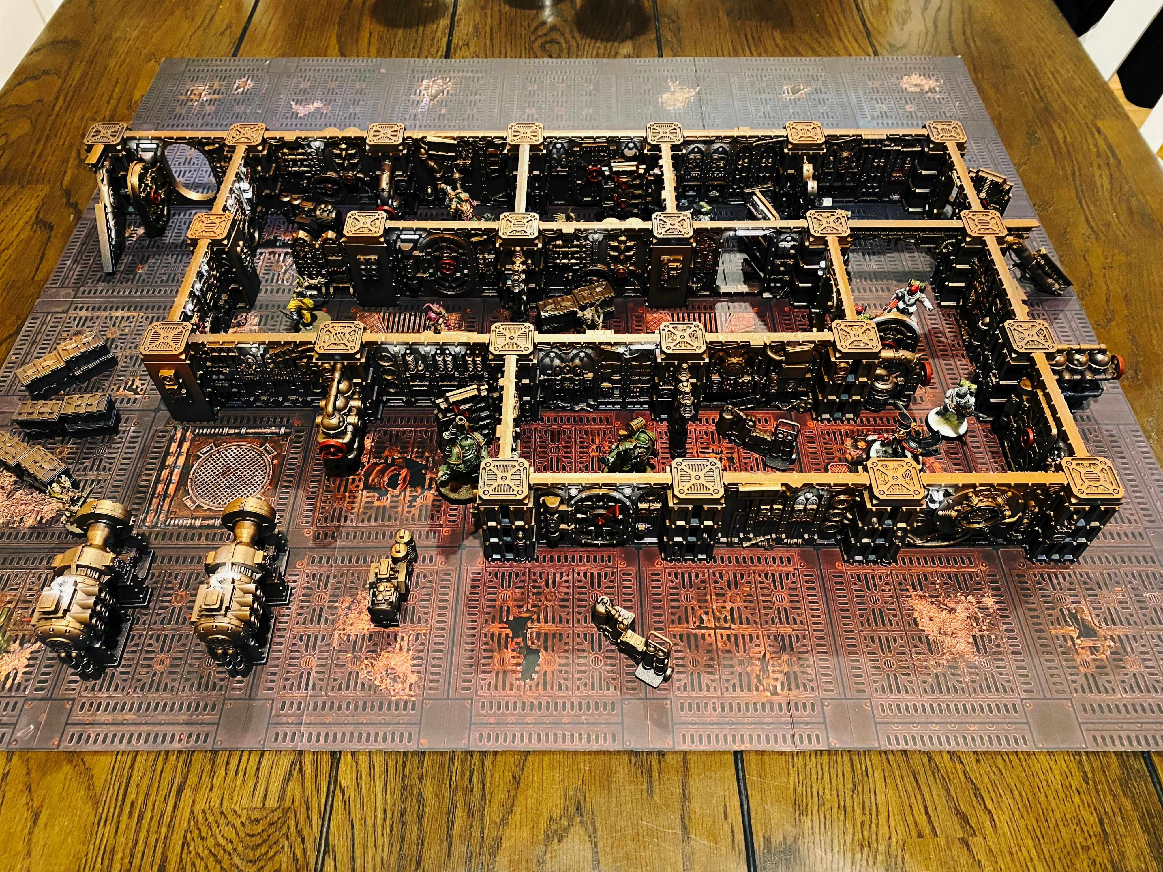 A photo of a bunch of steampunk-looking terrain on a game board, it's all walls and put together to look like the interior of a cramped gothic spaceship. There's miniatures in it, vile Plague Marines and Poxwalkers approaching through the corridors from the left, facing off again several female warriors in light blue/grey armour and wielding various guns.
