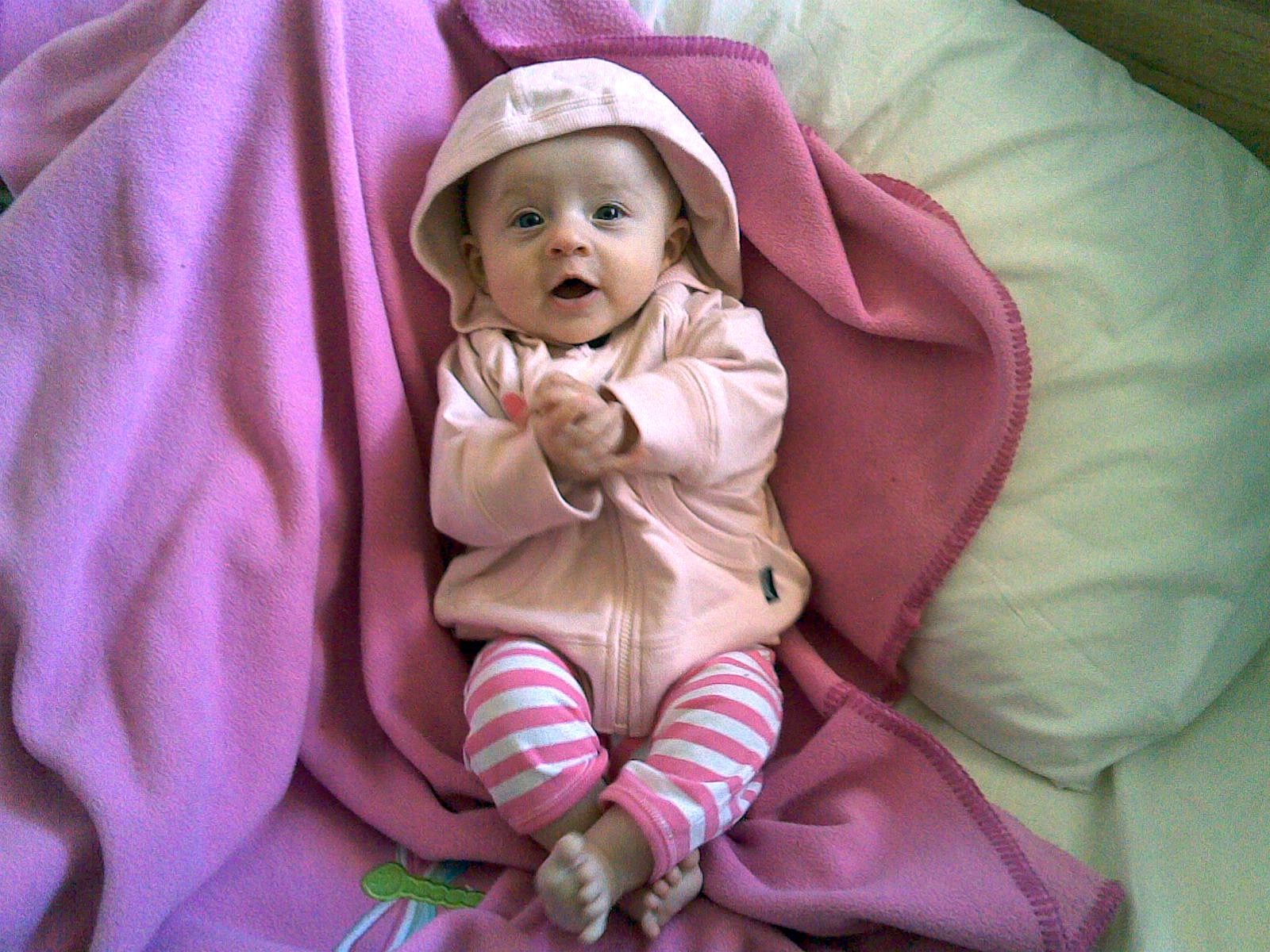 A photo of a small baby lying on a pink blanket with pink and white stripy pants on, and a pink hoodie. She has her mouth open in a big O shape and is mid-flailing her arms around.