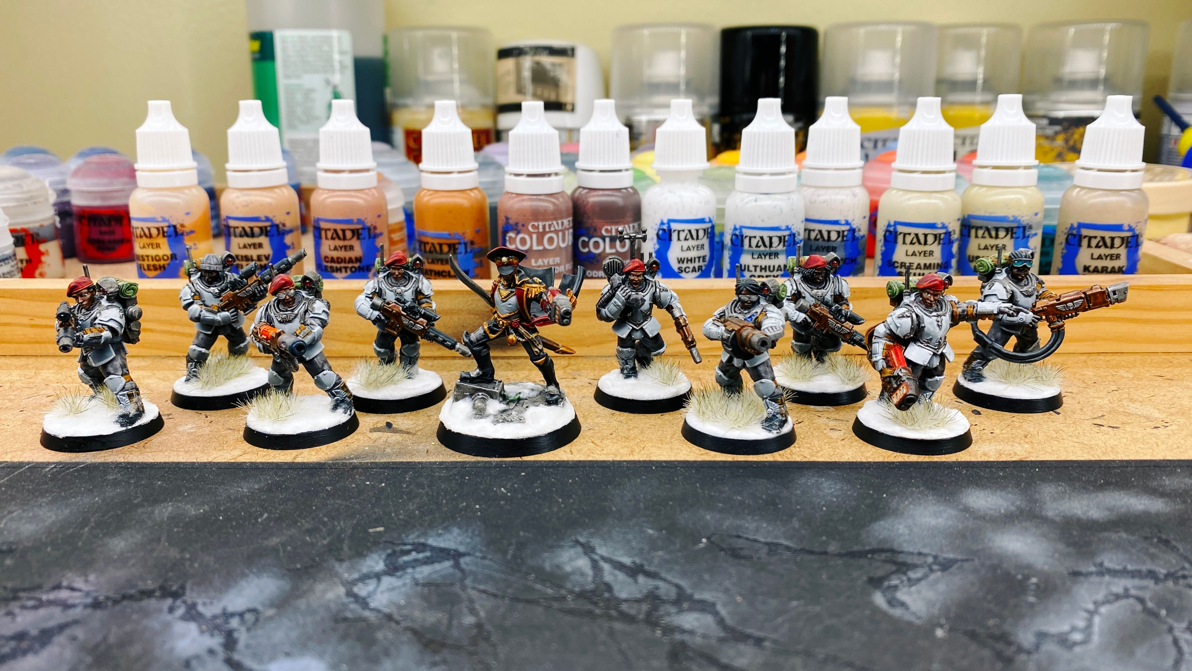 A photo of the full squad of Astra Militarum Tempestus Scions. In addition to the leader in the previous photos, there's another nine, they're all wearing big blue-grey armour and are wielding various guns, they've all got black or brown skin, and they all look extra bad-ass because they're wearing red berets and a couple of them have sunglasses on. The bases are done so they all look like they're standing in snow.