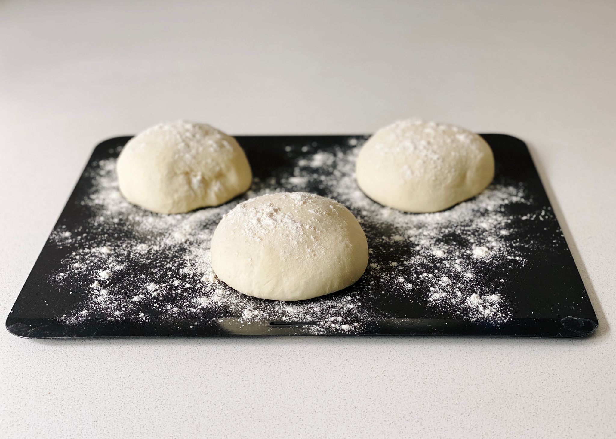 A photo of three round pizza dough balls sitting on a floured baking tray.