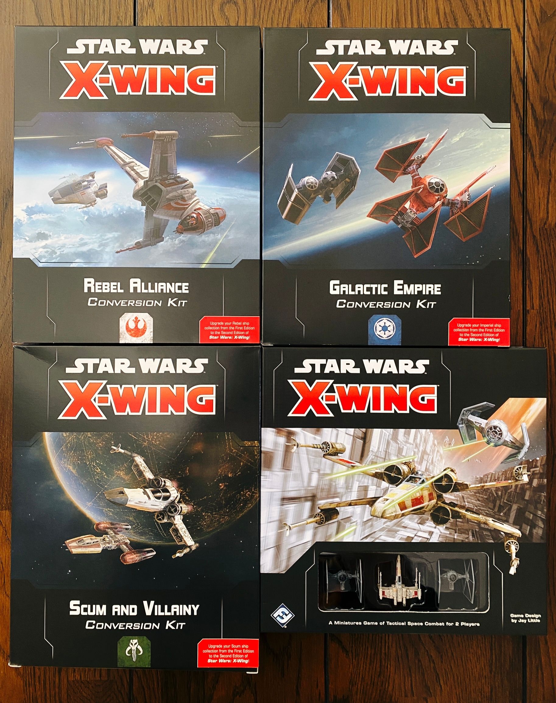 A photo of four boxes for the Star Wars miniatures game "X-wing". One is the 2nd Edition core set which has two TIE Fighters and one X-wing miniature in it. The other three boxes are conversion kits for the Rebel Alliance, Galactic Empire, and Scum and Villainy factions that contain all the tokens and cards and other things needed to use the ships you'd bought for 1st Edition with the new 2nd Edition rules.
