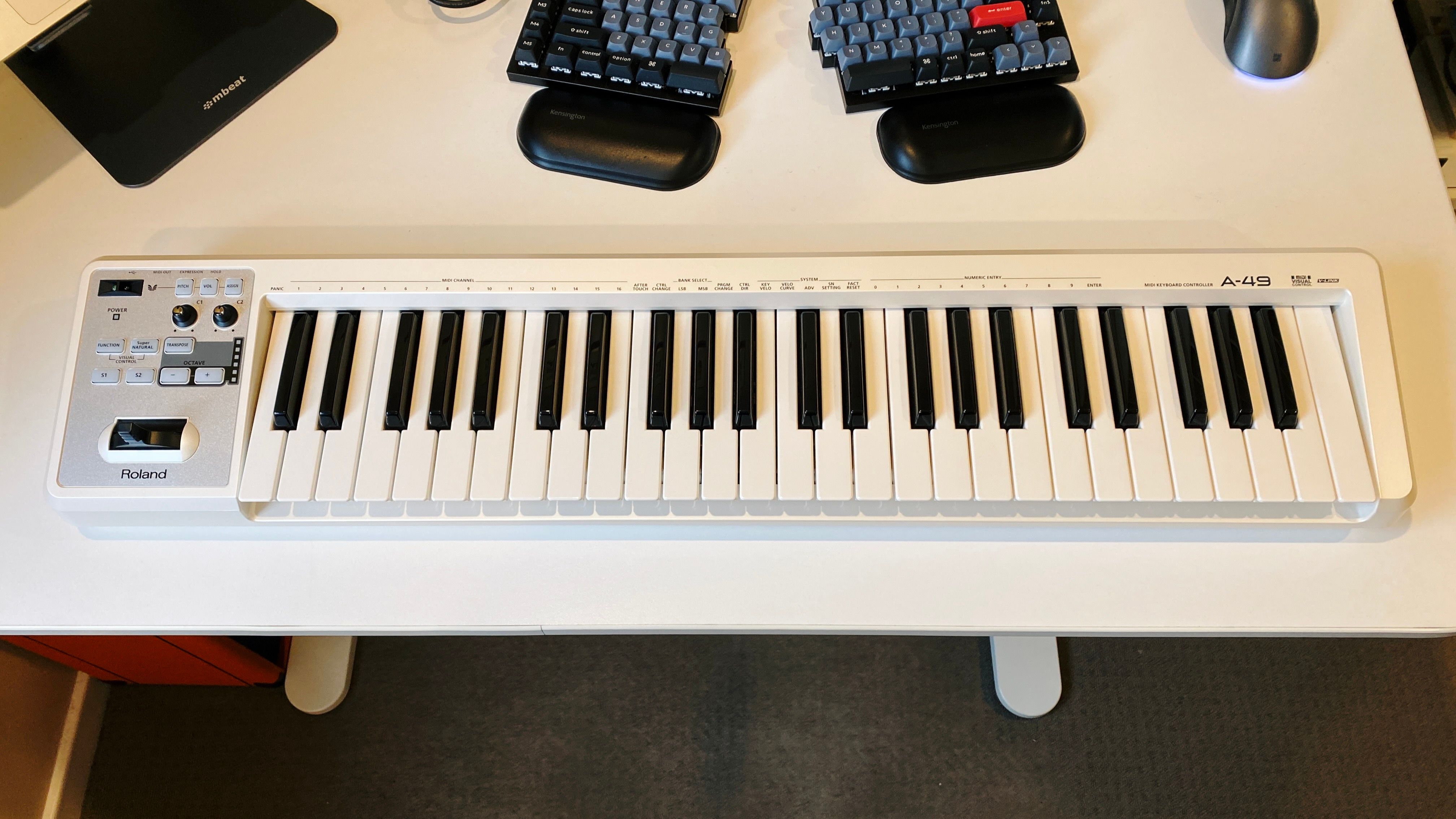 A photo of a Roland A-49 MIDI keyboard in white sitting on my (also white) desk. As the name suggests it has 49 keys, and has a number of buttons and knobs on the left hand side.