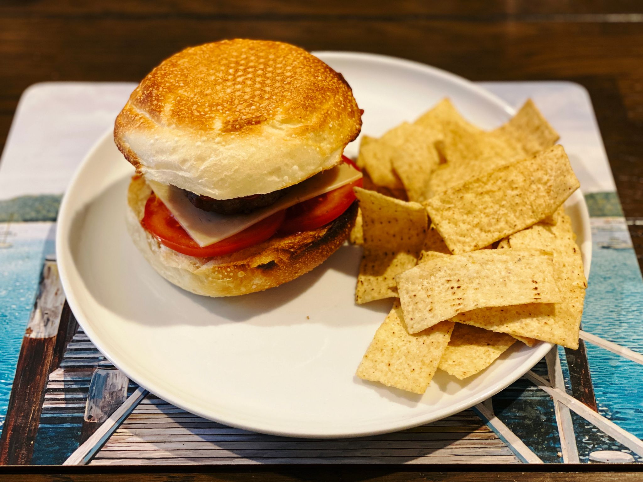 A photo of a home-made burger with tomato and cheese on it sitting next to a pile of corn chips on a plate.