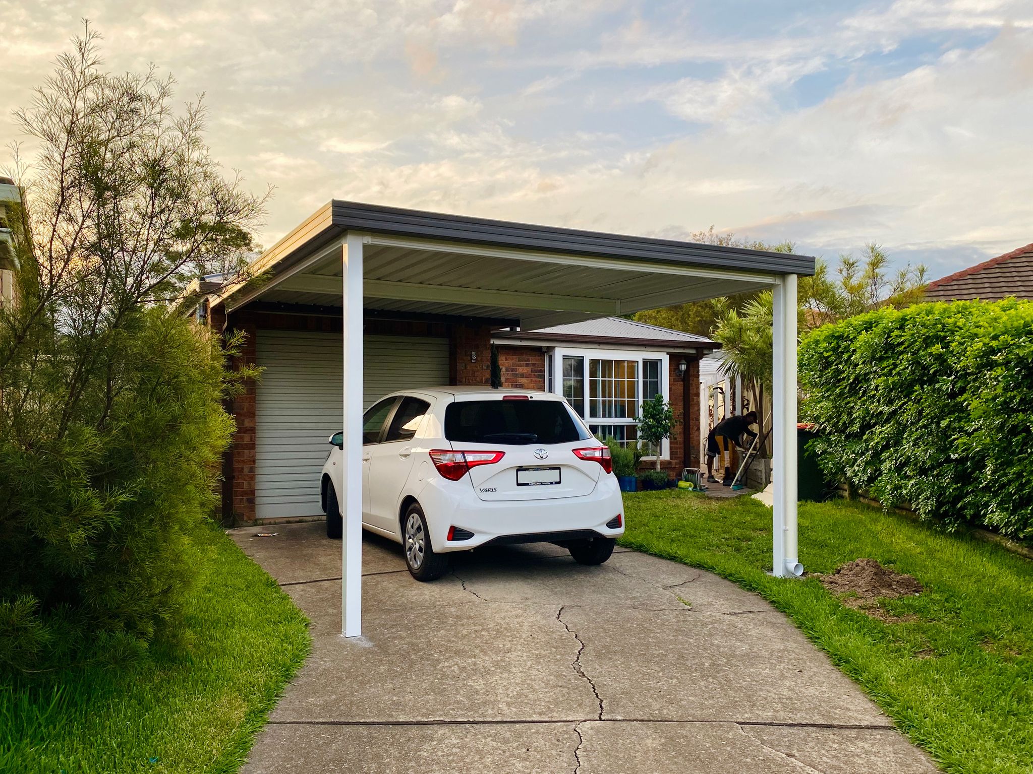 A photo of a carport with dark grey guttering and a white roof and posts, with a white Toyota Yaris parked under it. The front left post is partially on the driveway and the front right one is over on the grass due to the shape of the driveway.