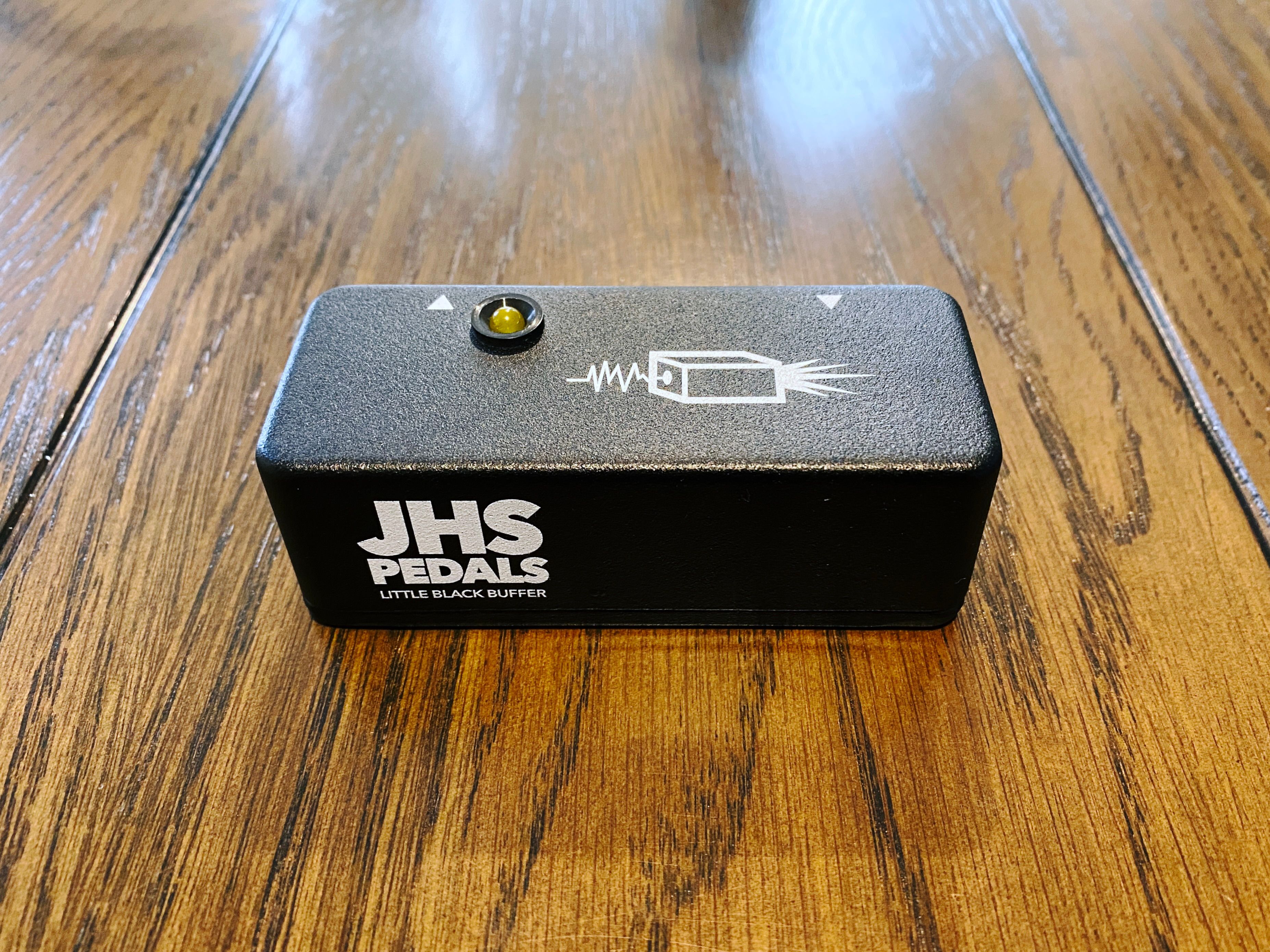 A photo of a little rectangular black box that says on the side of it "JHS Pedals" and underneath "Little Black Buffer". There's a single yellow LED on the top of it (the input and output plugs are on the very back and not visible in the photo).
