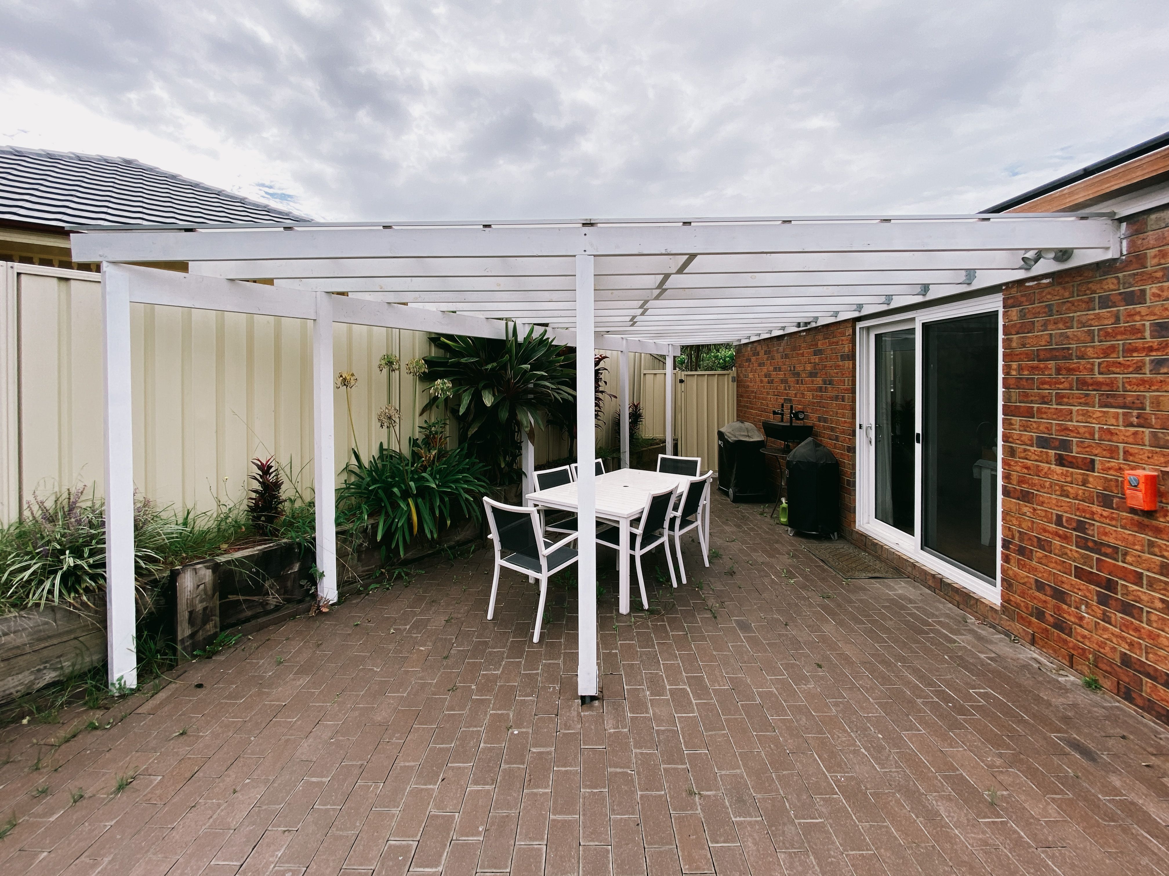 A photo of our old pergola. It's done in timber with white paint over it, and the roofing is polycarbonate. There's a vertical post in the middle of the longest span and the whole thing looks a bit shit.