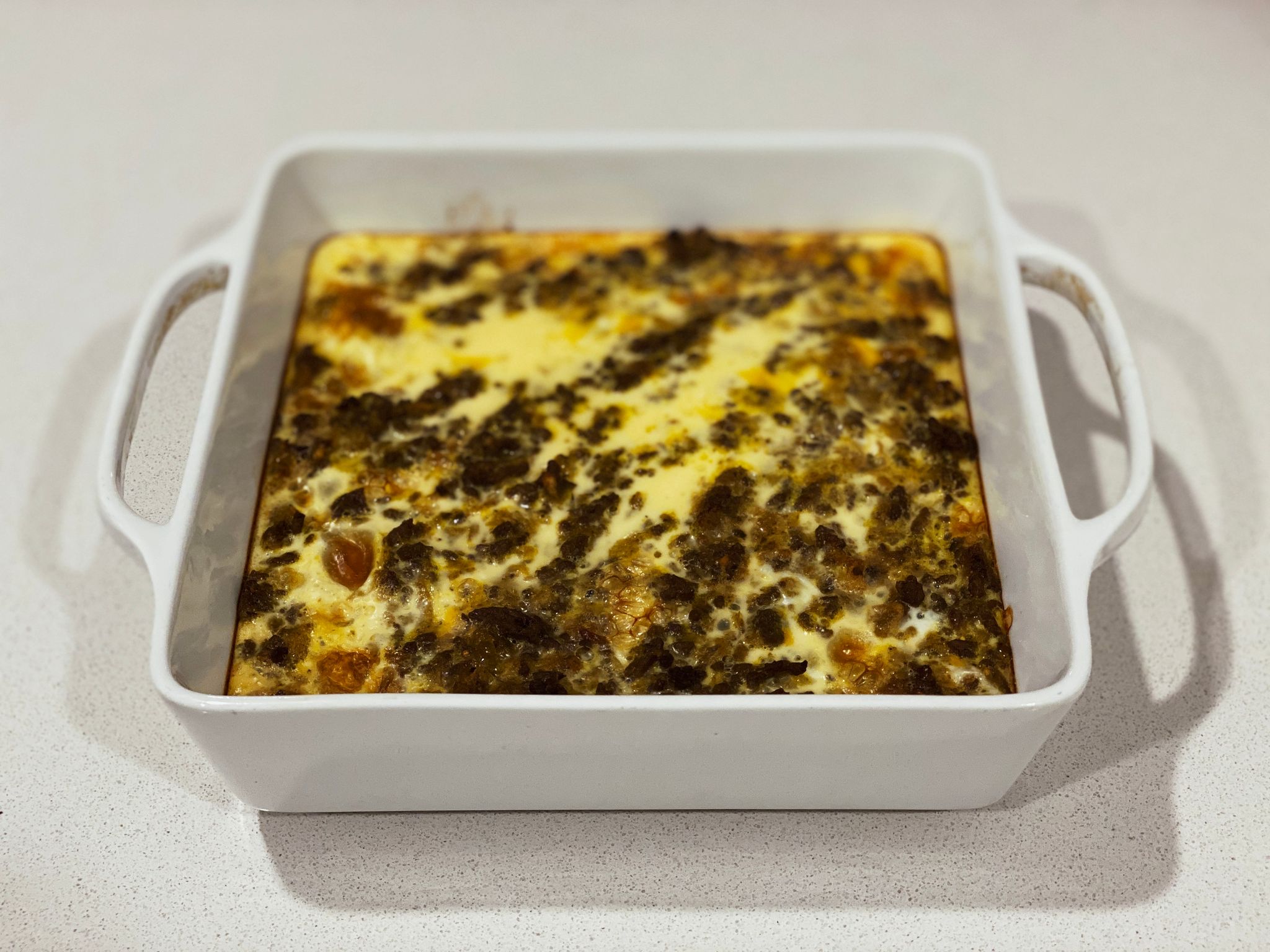 A photo of a square white ceramic oven dish with food in it, beef mince is visible popping out the top of the whisked egg that's sitting on top of it all.