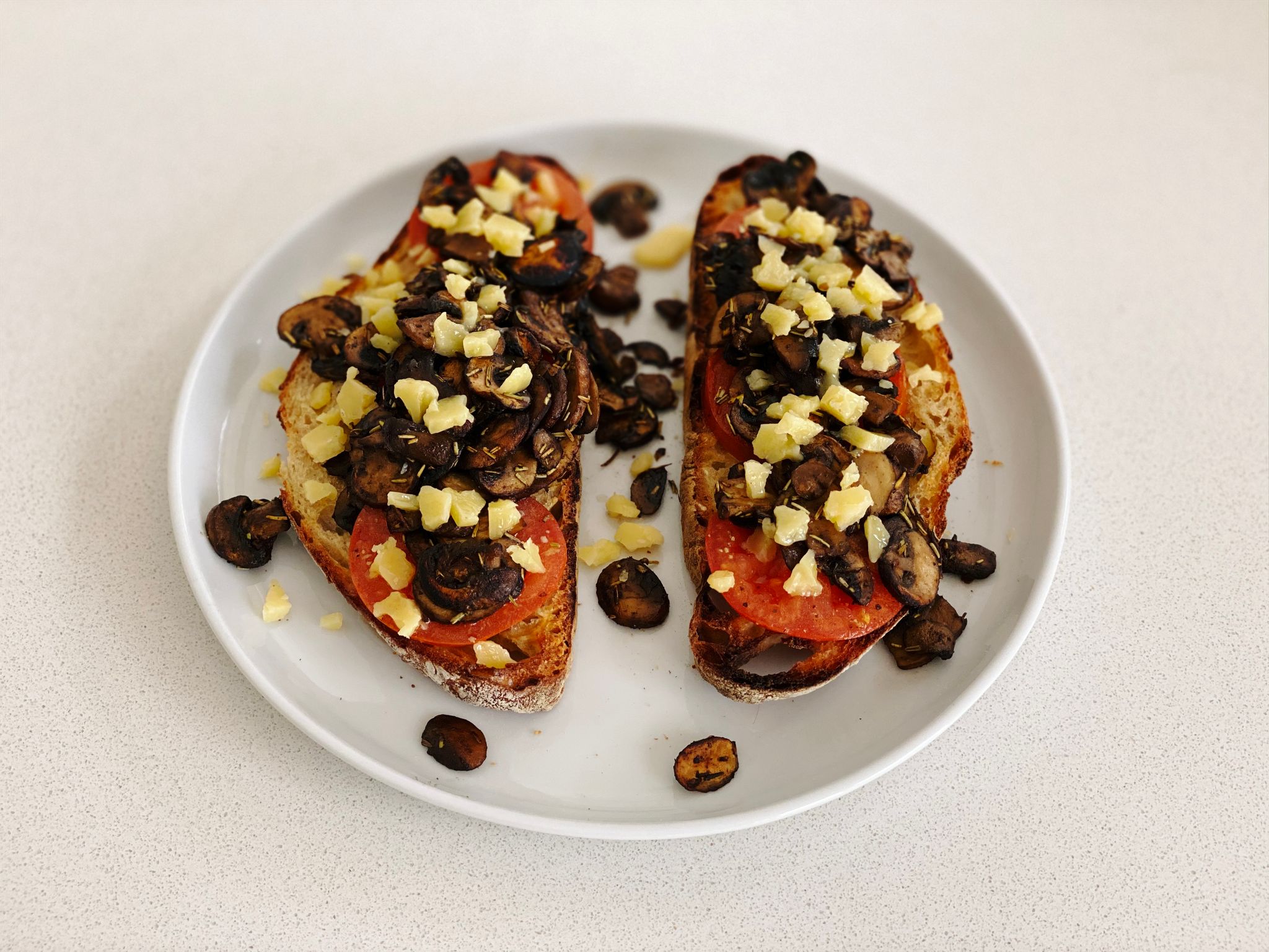A photo of two pieces of toast sitting on a plate, with cooked tomato and mushrooms on top, and crumbled cheddar cheese on top of that.