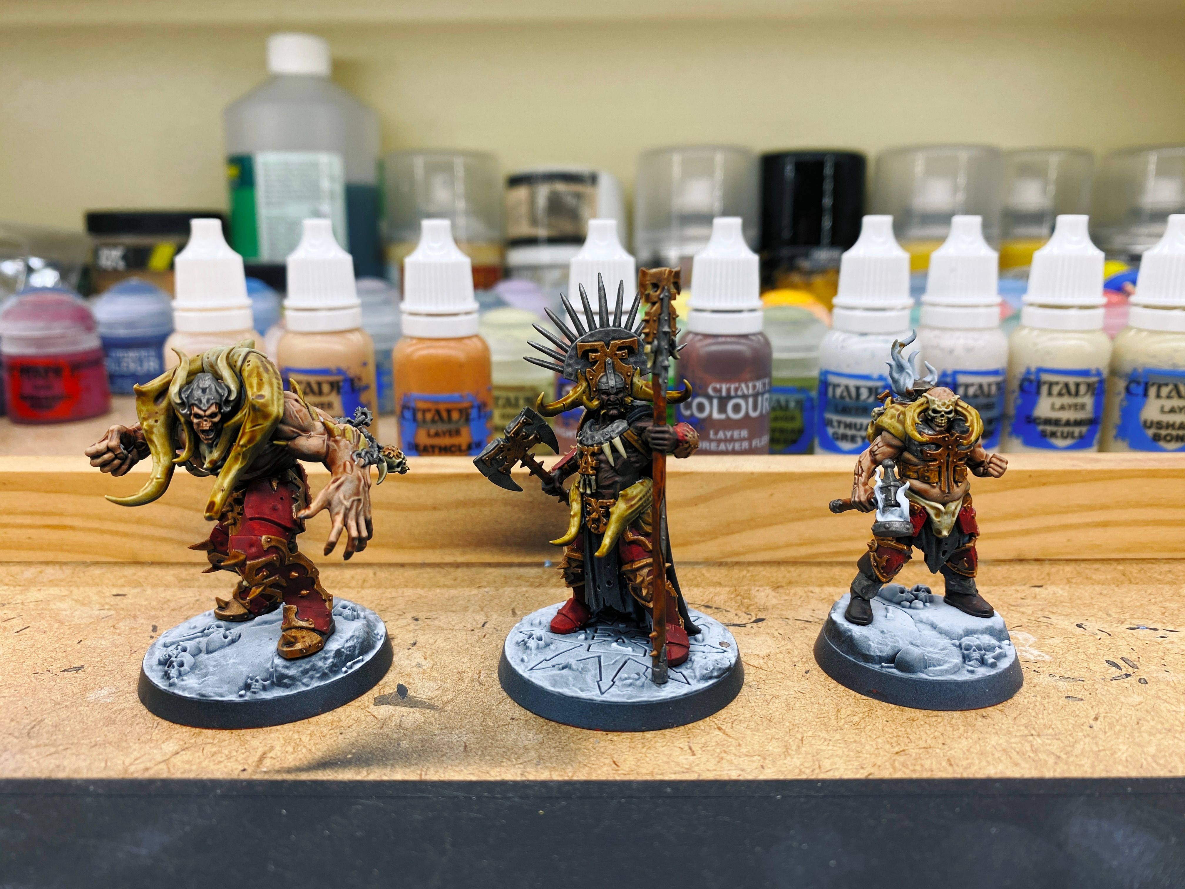 A photo of the three miniatures that make up the Warhammer Underworlds warband "Gorechosen of Dromm". They're three beefcake dudes, one's a Black guy with a big ceremonial headdress on and is wielding an axe and a staff, one has very Immortan Joe from Max Mad vibes but also with rocking dad-bod, and the other is part demon with absurdly large arms and big horns over his shoulders.