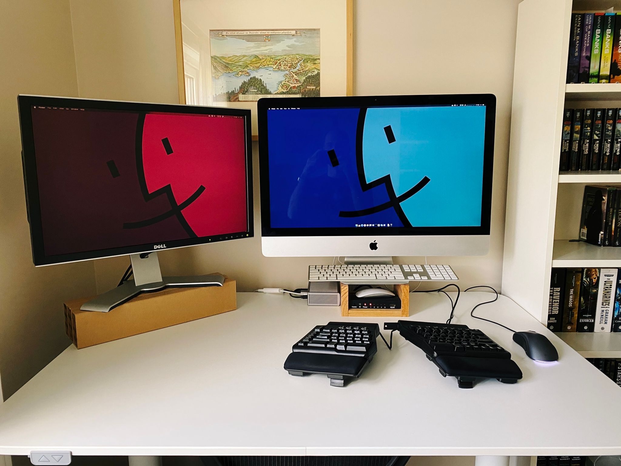 A photo of my old computer setup: a 27" 2013 iMac in the middle sitting atop a slightly-dodgy home-made wooden stand, with a 15 year-old 24" Dell to the left of it sitting on an even dodgier stand that consists of hard rectangular pieces of cardboard that were part of the packaging for something or other. There's a CalDigit dock next to the iMac with a ton of cables coming out of it.