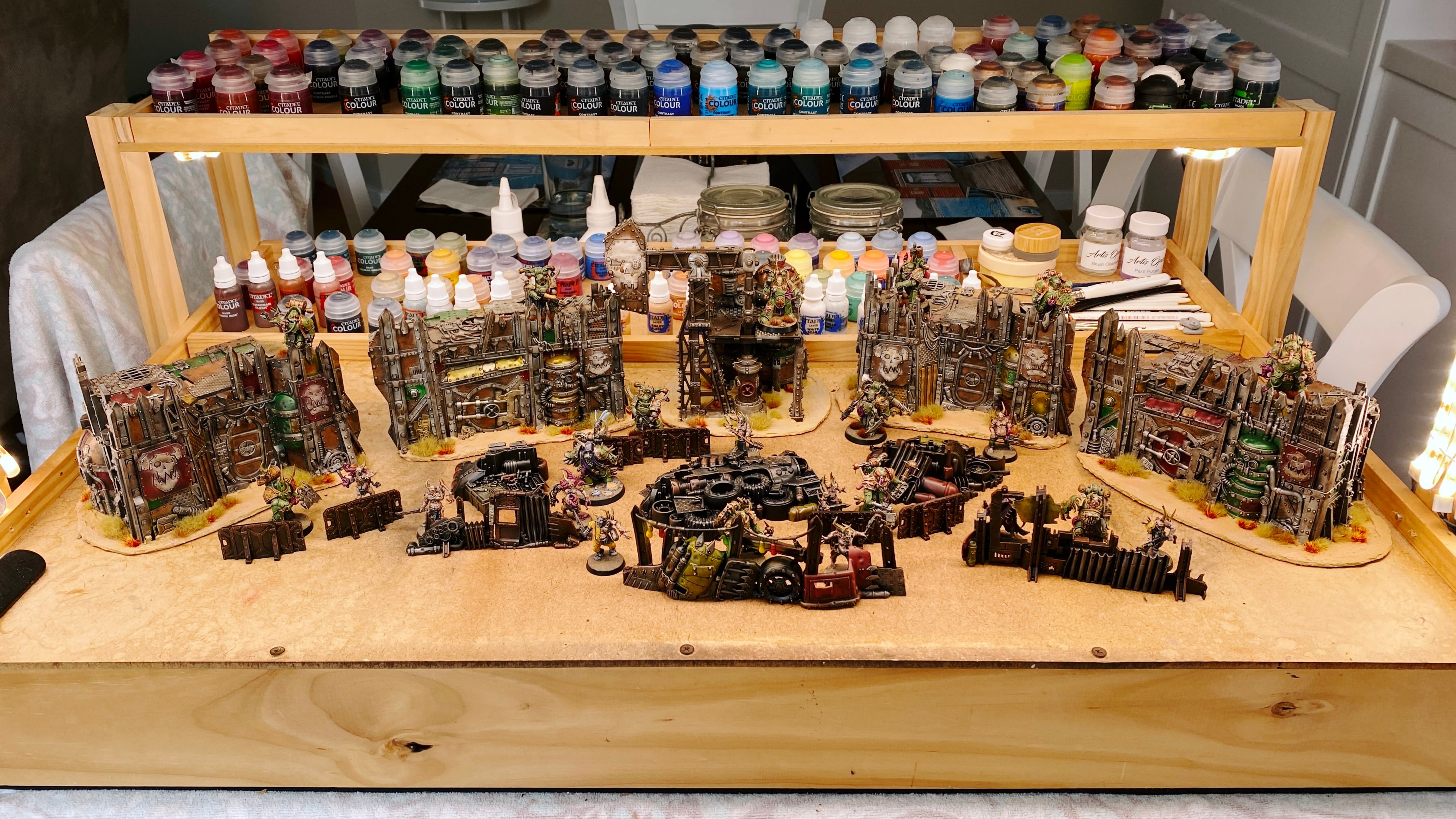 A photo of entire set of EXTREMELY ramshackle pieces of terrain from the Warhammer 40,000 Kill Team: Octarius box from Games Workshop. At the back are four parts of an Ork fort that look like they belong in a junk yard, the walls and platform on top are made up of a whole ton of pieces of metal scrap, like big plates, corrugated pieces, massive beams, there's a door in there, and a couple of empty gas cylinders. There's also an old rig thing with the big pumping arm in the same ramshackle style, and scattered in front of these larger pieces are piles of scrap metal and chest-high walls also made out of all sorts of pieces of scrap.

All the big pieces are on cut-out pieces of cardboard that are covered in a texture paint that makes it look like sand, and there's various tufts of dry grasses growing around the base of the walls.
