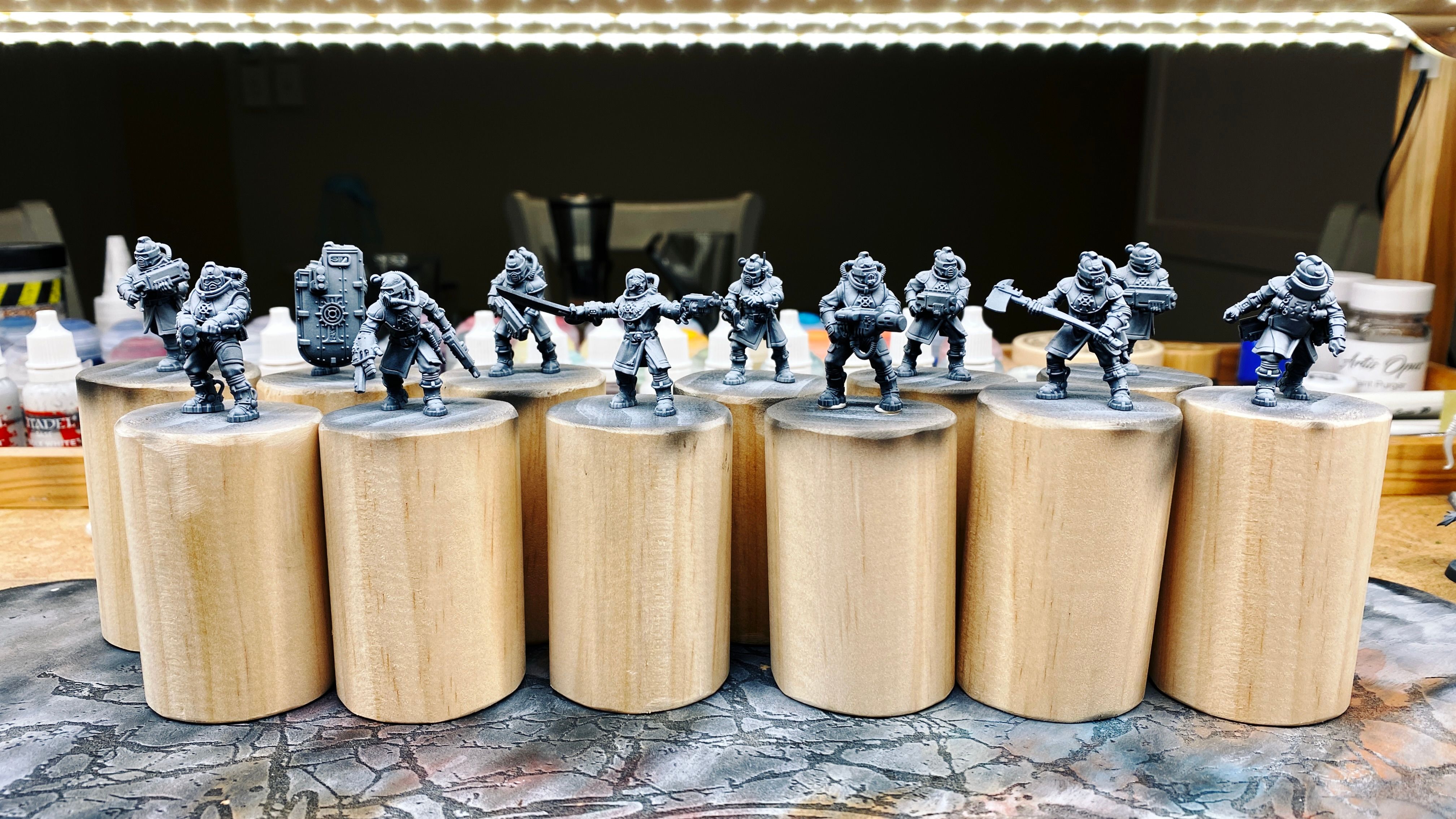 A photo of twelve armed and armoured human soldier miniatures assembled and undercoated. Apart from the leader who is (as is traditional in the Warhammer 40,000 universe) not wearing a helmet, all the rest have cool-looking helmets on that are very reminiscent of old diving bell helmets, just with a regular visor instead of a narrow round window. They all have tanks on their backs and pipes leading from them to the masks, and have a variety of guns. One of them has a huge riot-shield-type thing and is aiming a futuristic shotgun through a slot in the top of it.