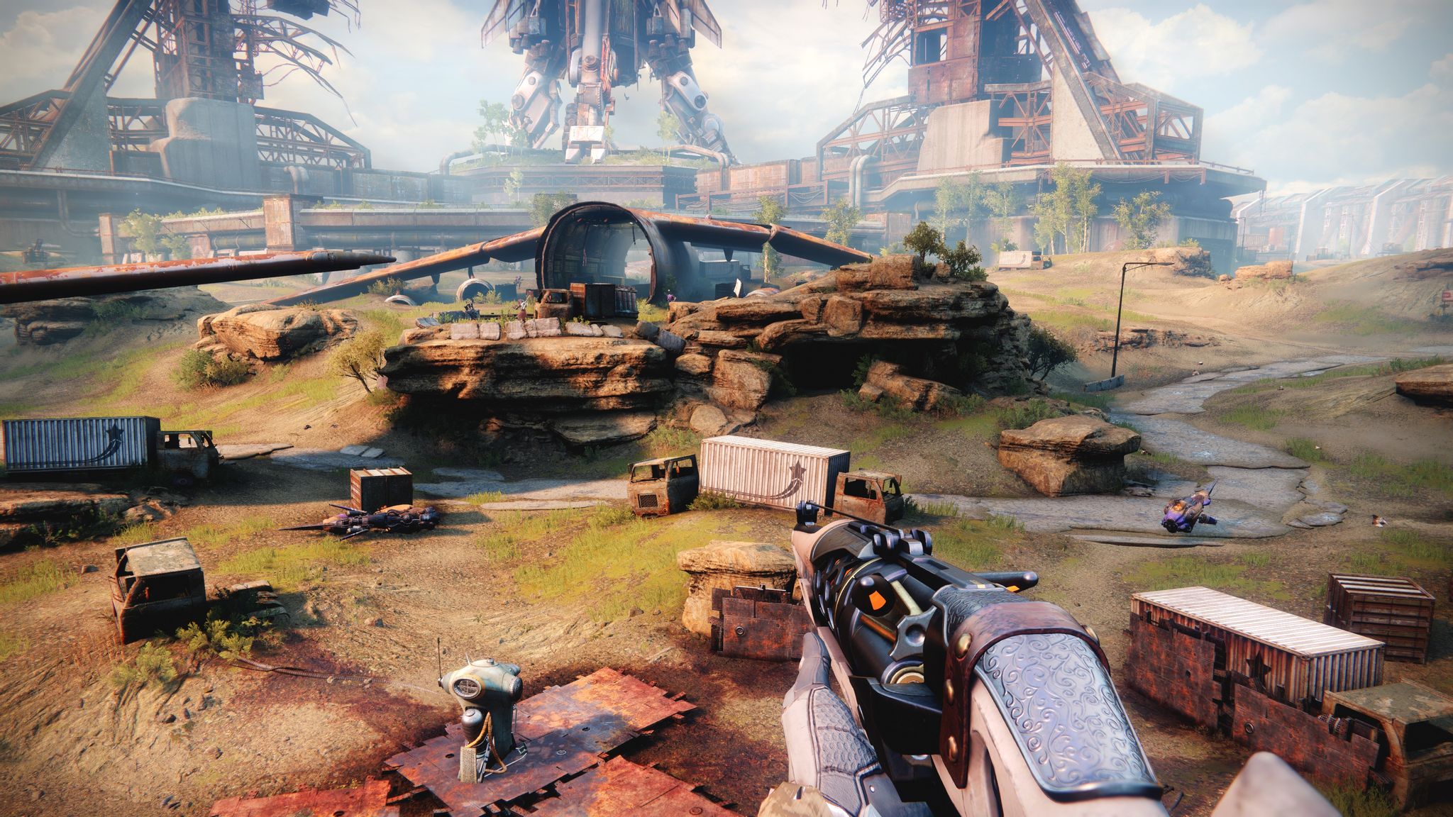 A screenshot from Destiny 2 showing a bunch of rusting metal boxes and a hollowed out shell of a large plane in the distance. The ground is fairly rocky and barren but has more grass and greenery than it used to in Destiny 1.