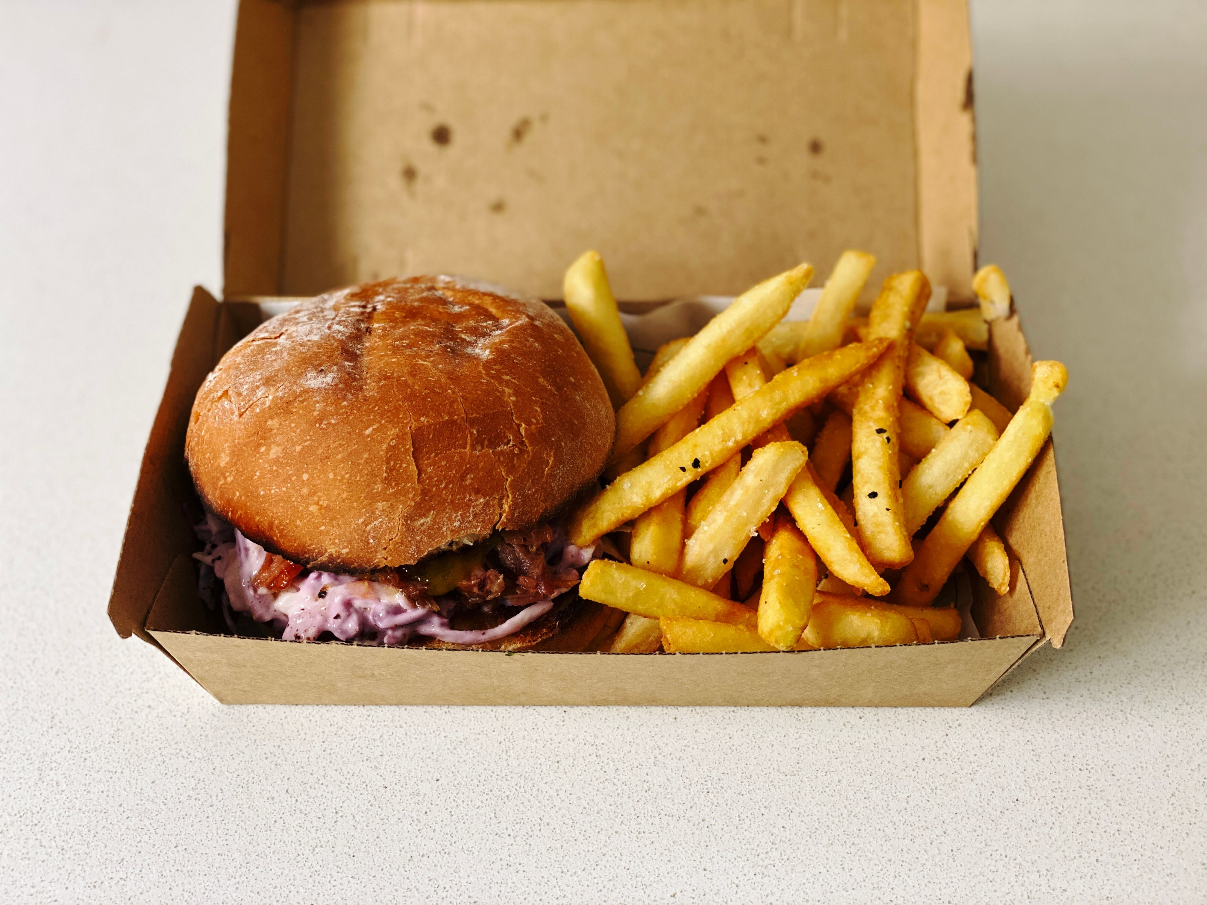 A photo of a pulled ham burger with slaw and a hot chips in a brown cardboard takeaway box.