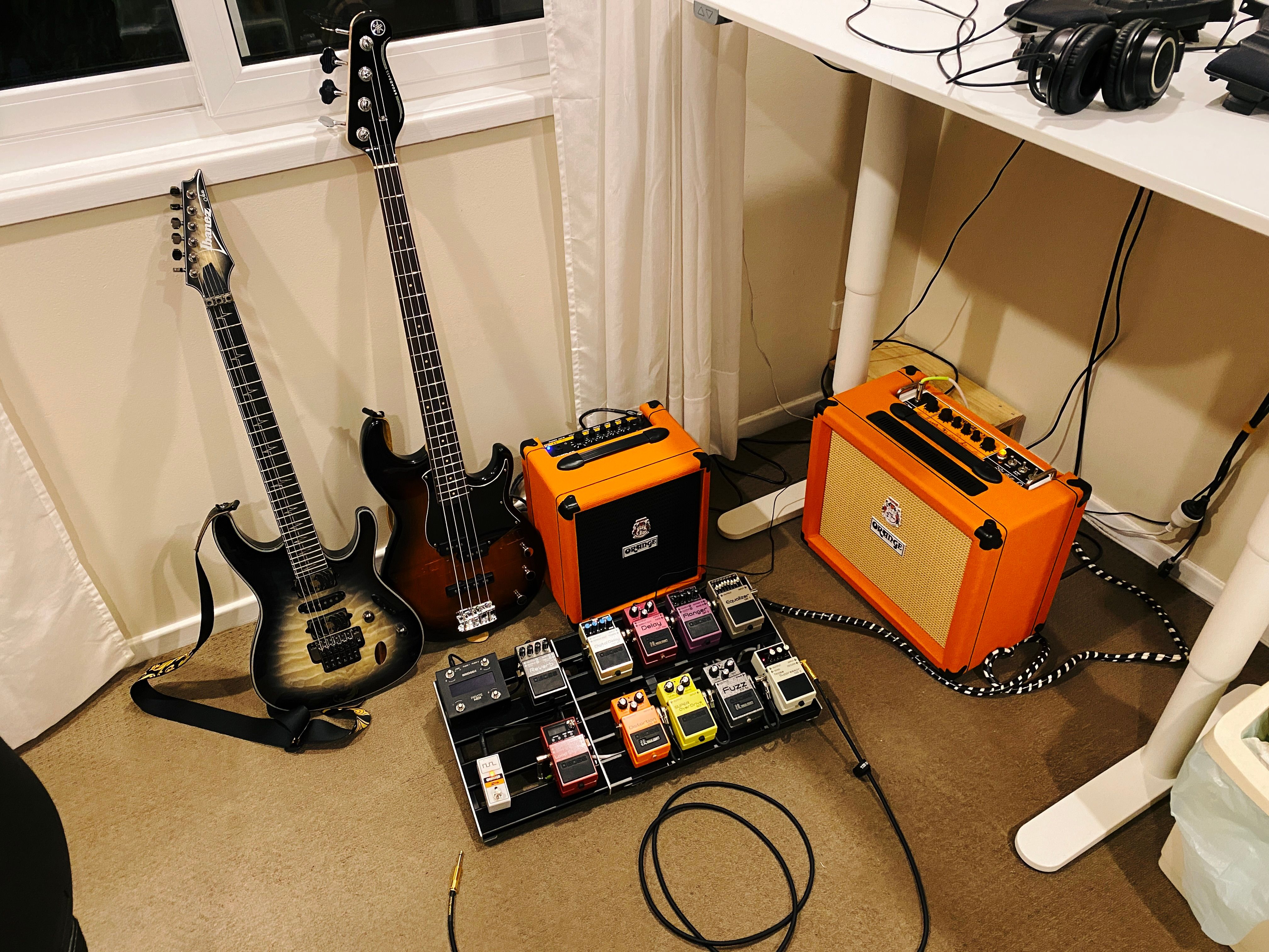 A photo of an electric guitar and bass guitar propped against a wall, sitting next to a bass guitar amp and a regular guitar amp, with a pedal board filled with pedals sitting in front of the whole lot, and cables going everywhere.