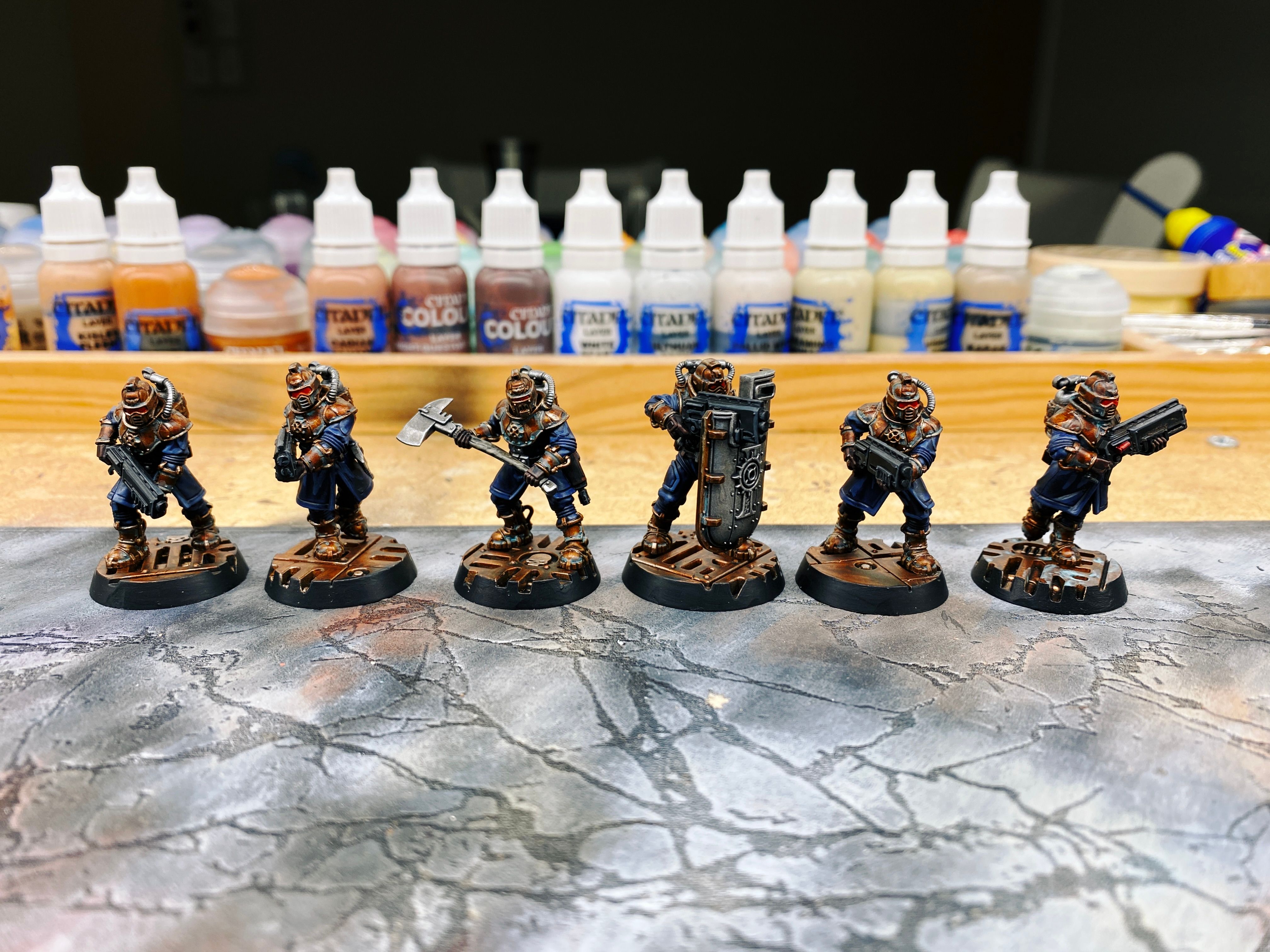 A photo of six armoured human warrior miniatures. Their armour is a deep brass colour, they're wearing big mag-boots of the same colour, and their clothes are a rich dark blue. The helmets are reminiscent of diving bells except with a cool-looking visors instead of a round viewing port. Four of them have big short guns, one is wielding a huge axe, and the other has a massive shield that's taller than he is, with a viewing port and a slot for his gun to sit in so he can see and shoot while remaining behind the shield.