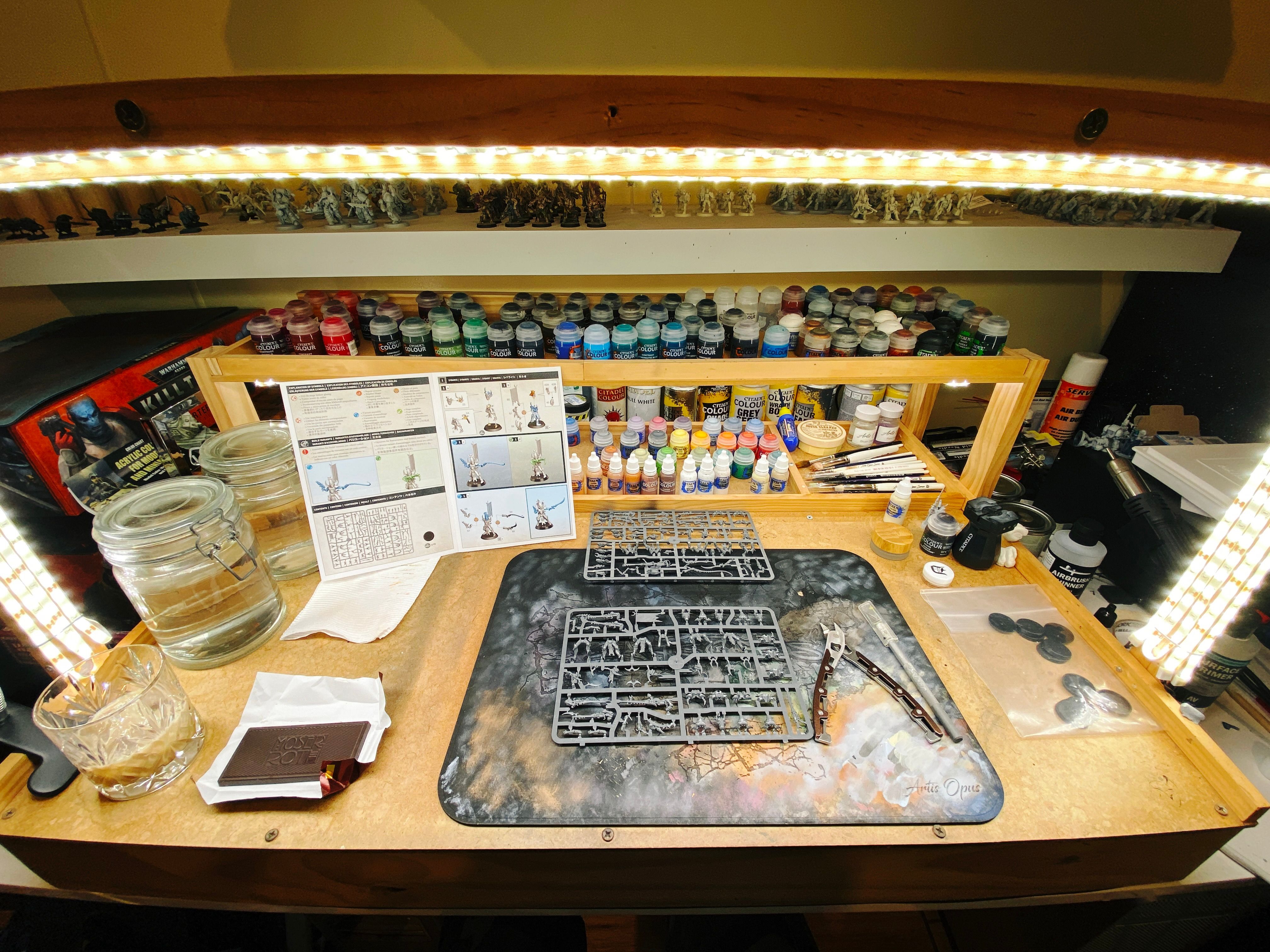 A photo of my painting desk with a two sprues of miniature parts on it, ready for assembly. Also a piece of dark chocolate and an alcoholic beverage.