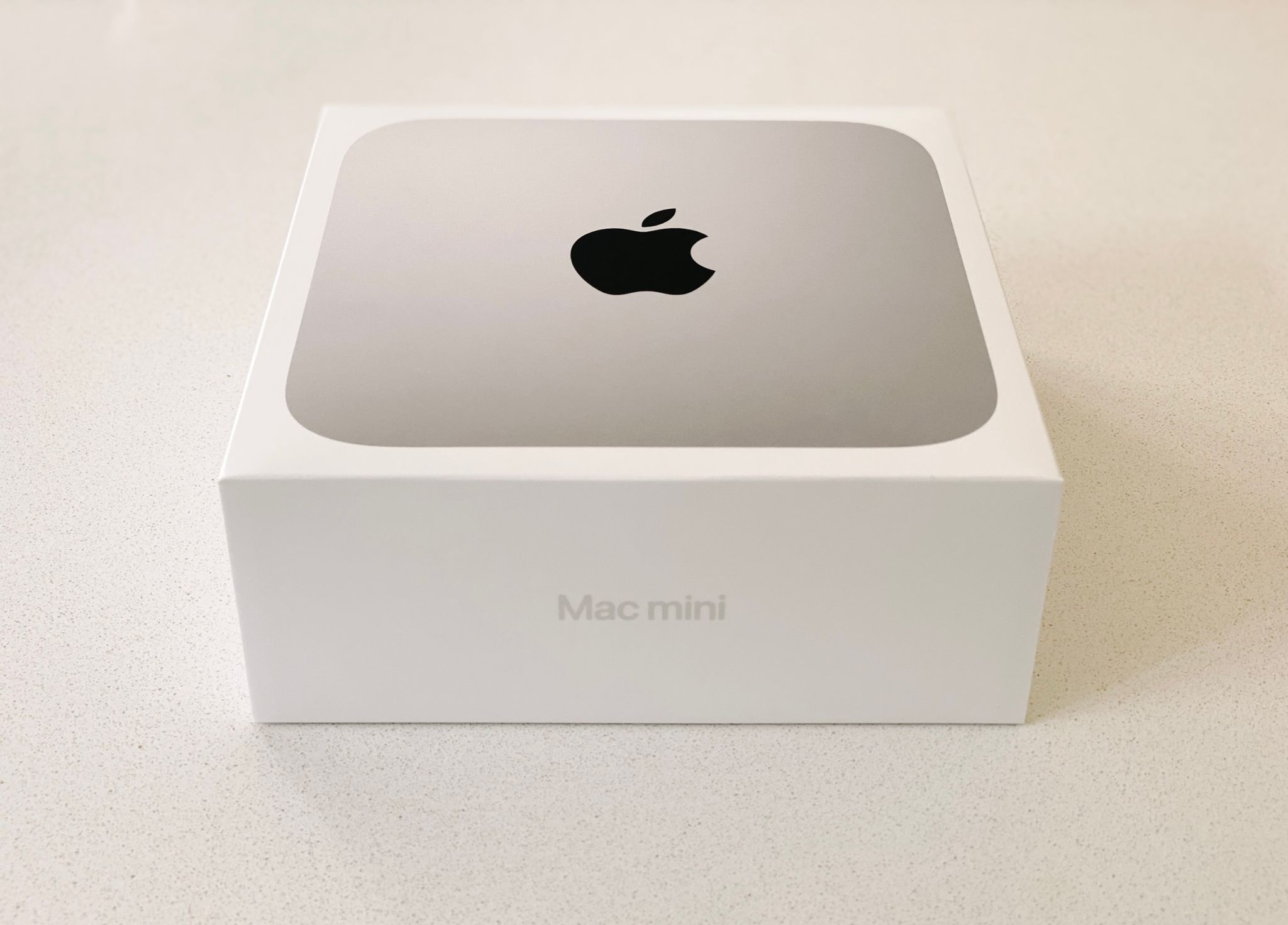 A photo of the box of a Mac mini. It's white and very minmal, with "Mac mini" written in silver on the front and a photo of the top of the Mac mini itself on the top of the box.