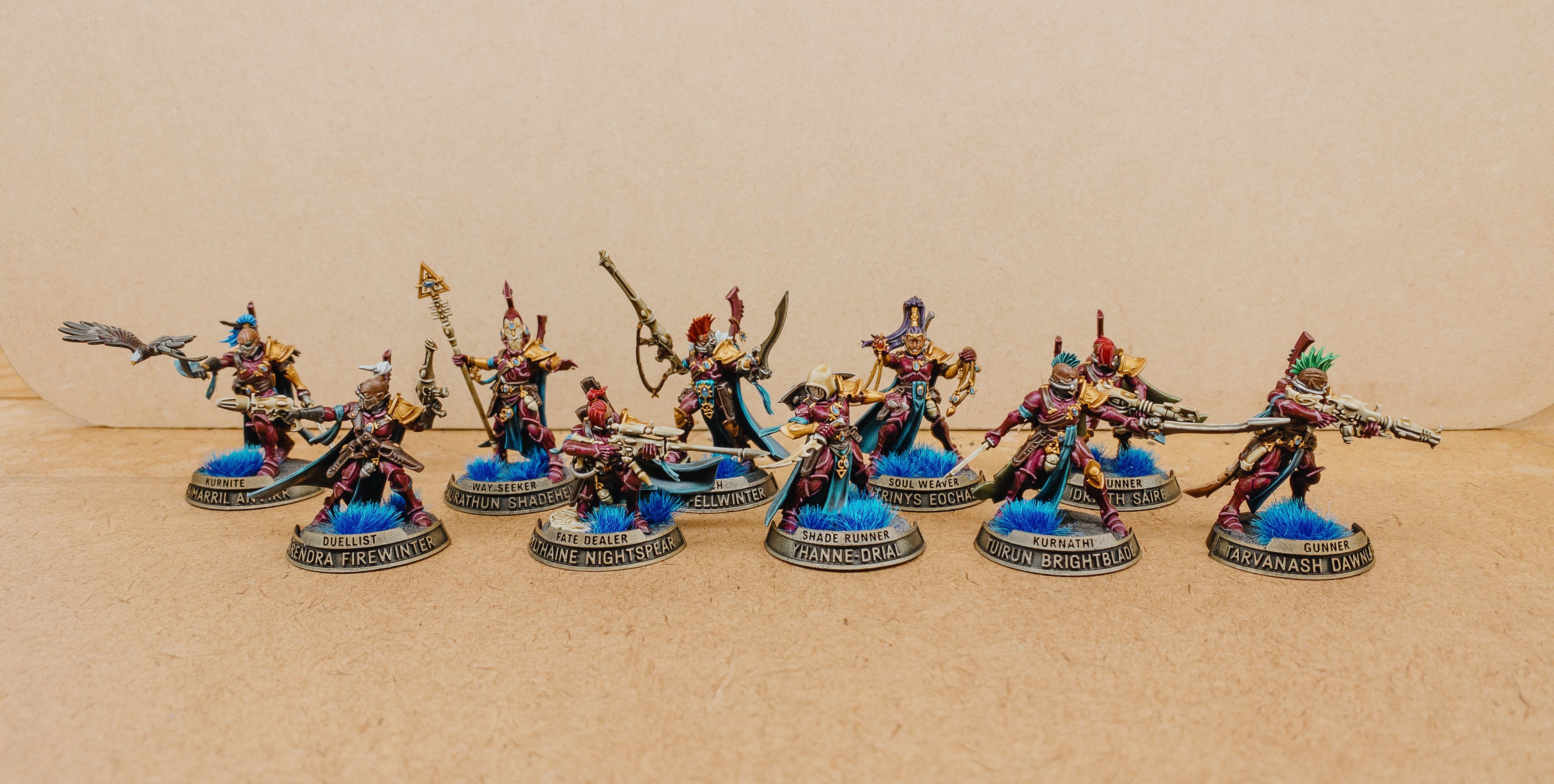 A photo of the ten miniatures that are my Corsair Voidscarred squad. They're essentially space elf pirates, all their armour is elegant and flowing and a rich burgundy colour, and they have cool turquoise capes. They're branding various ranged and close-combat weapons, and several of them have brightly-coloured mohawks. They each have a personalised name plate attached to the front of their bases, with a smaller bit of text on top that gives the unit type ("Fate Dealer", "Soul Weaver", "Way Seeker", and other such cool-sounding things), and a larger piece of text along the bottom that gives the unit's actual name, which are all sort-of Celtic feeling.