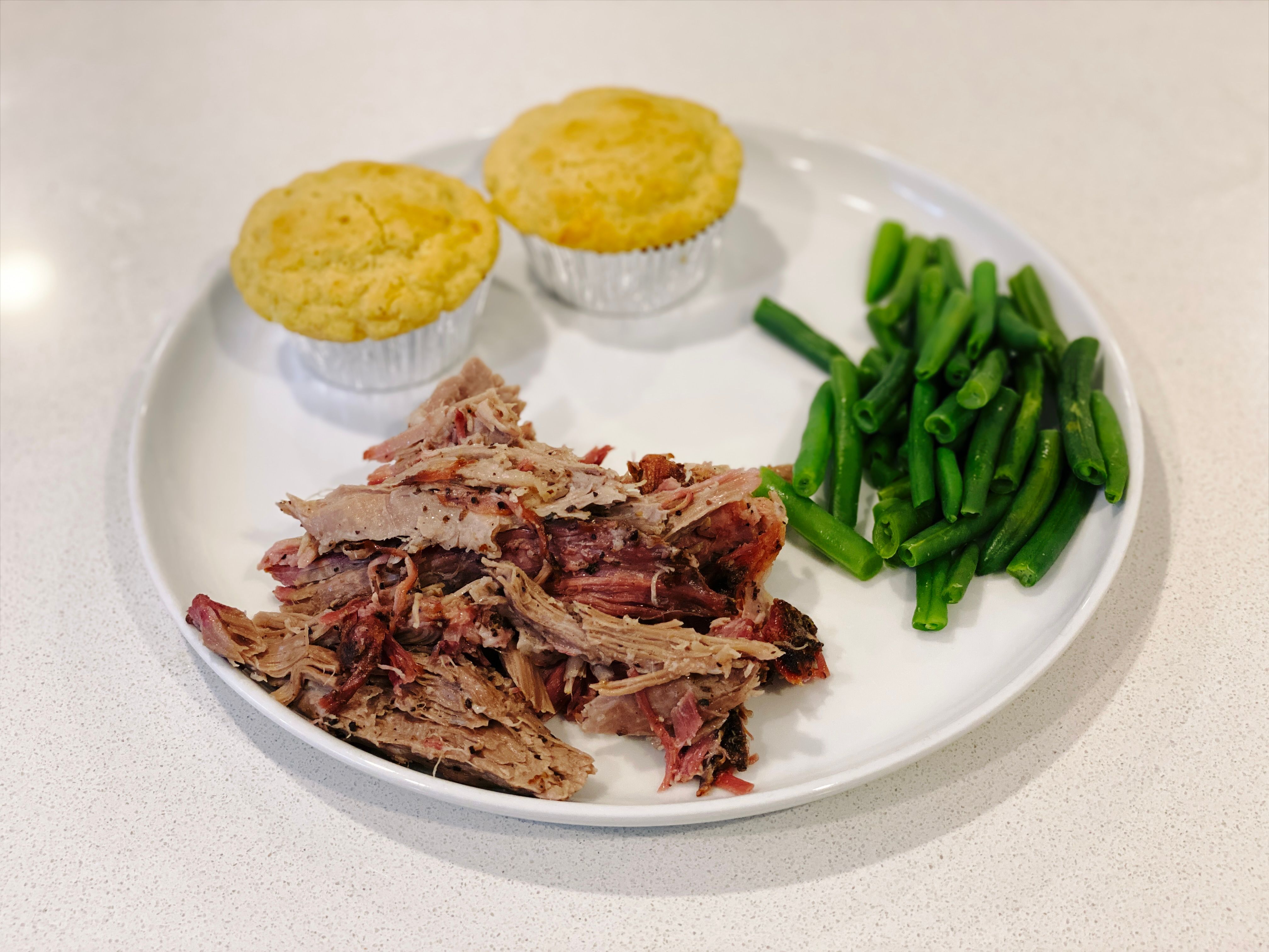 A serving of the pork collar butt on a dinner plate along with some green beans and two corn muffins.