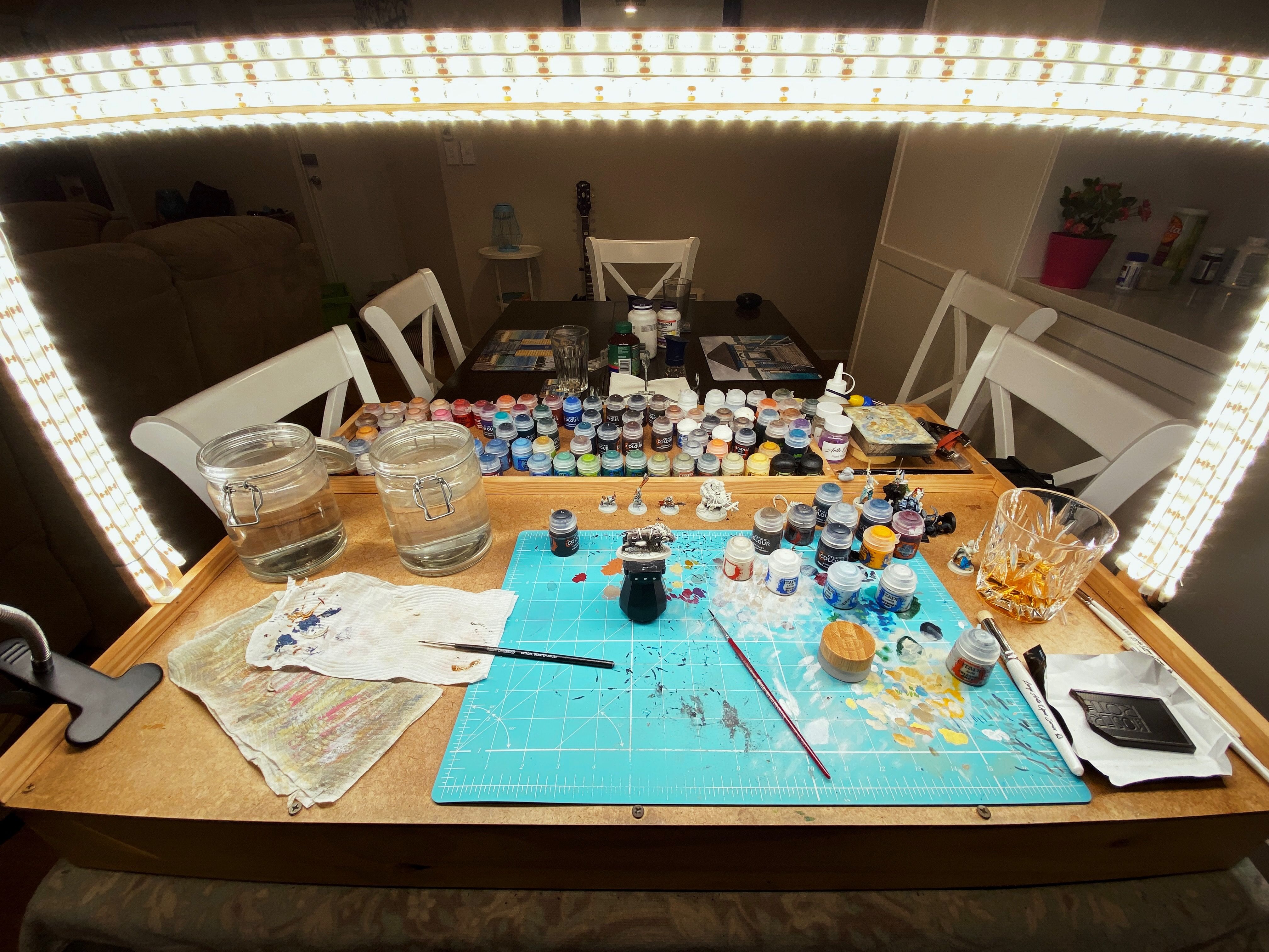 A photo of my DIY miniature painting table setup, it's made of wood and has a rectangular arch over it with a bunch of white LED strip lights attached to the inside of it. There's also two rows of the lights pointing directly forwards at the camera because of the aforementioned running out of room on the underside.