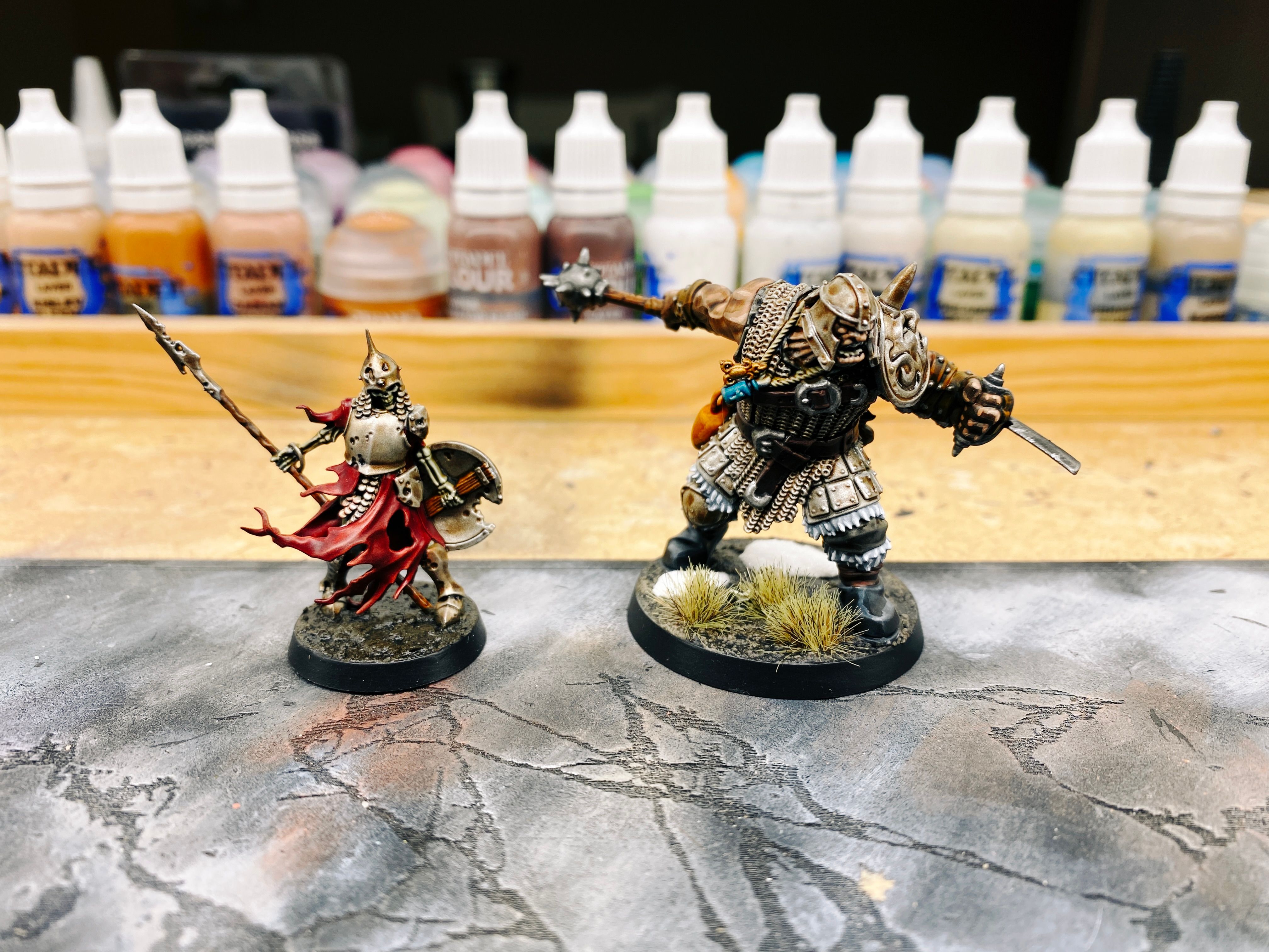 A photo of two miniatures side-by-side. One is a standard human-sized skeleton warrior, the other is an ogre who is head and shoulders taller than the skeleton. The ogre is wearing chainmail and a big helmet that leaves his face exposed, and has a big mace held out in one hand behind him like he's either about to hit something or has just finished doing a big backhand swing with it. In his other hand is clenched a big blade that's attached to a leather glove and that arm looks like he's about to stab someone with it.