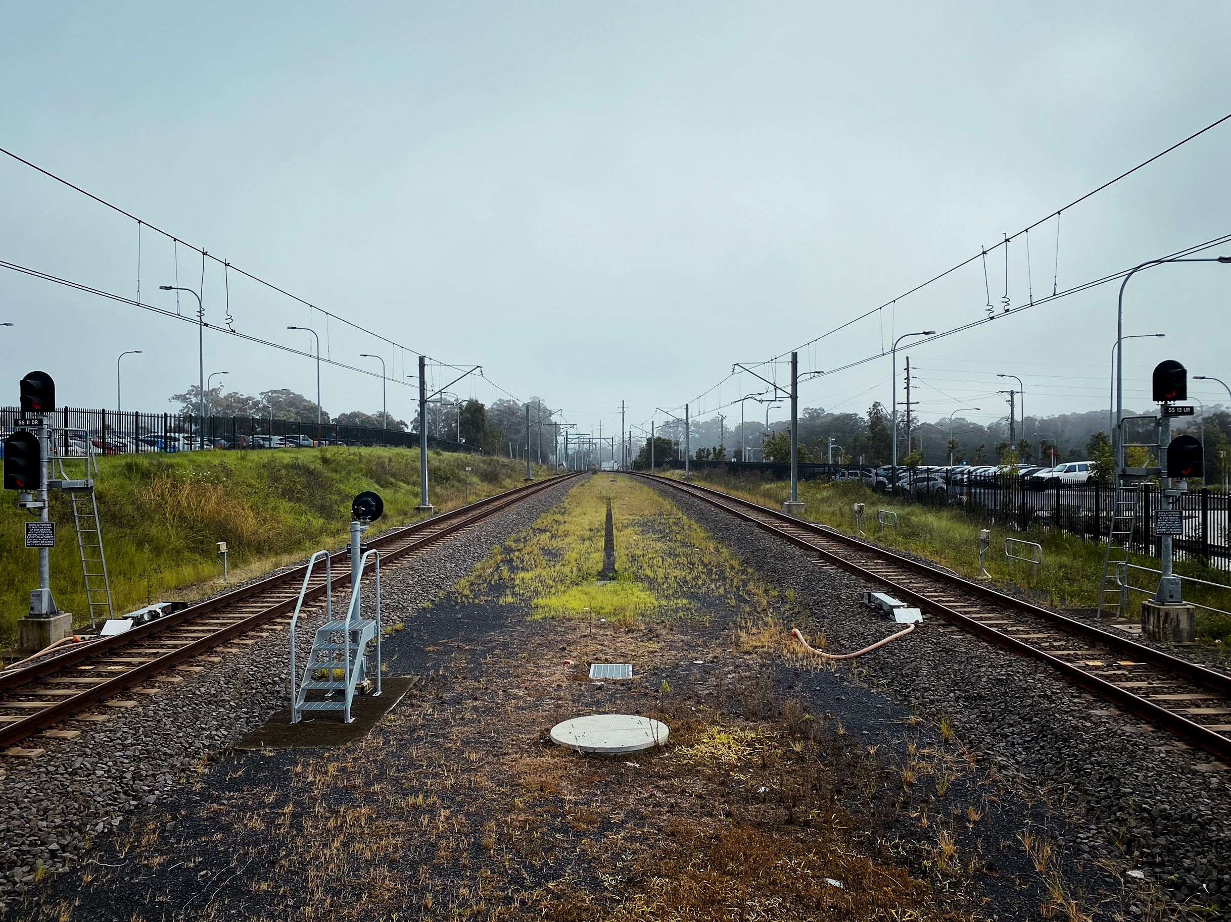 A photo taken from the end of a train station platform, looking down the tracks. It's overcast and slightly foggy, and the weeds between and on either side of the track are a little overgrown.