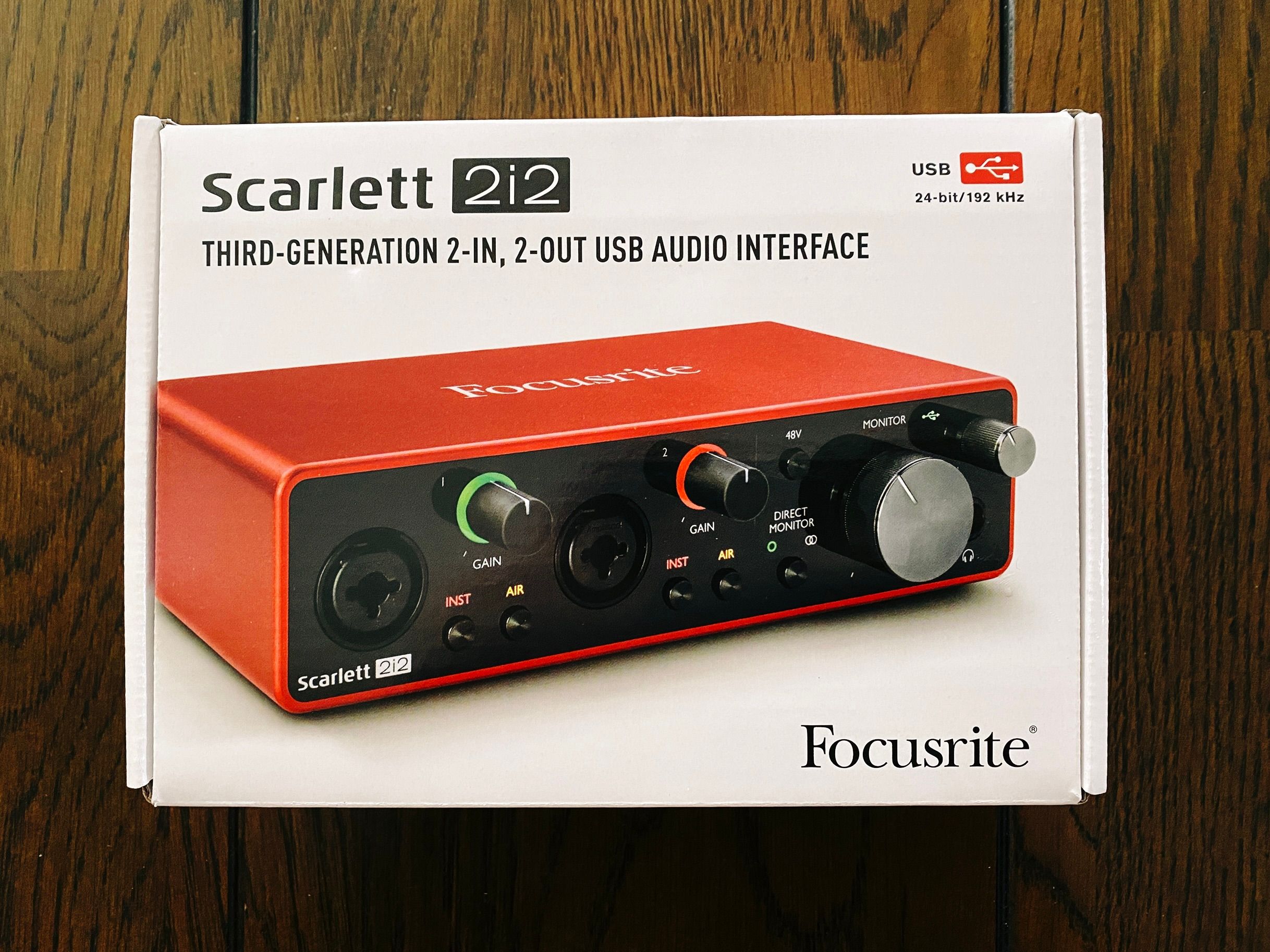 A photo of the box of a Focusrite Scarlett 2i2 Gen 3 audio interface. The top and sides of the interface are a rich deep red metallic colour, and on the front is shiny black with all sorts of dials and sockets on it.