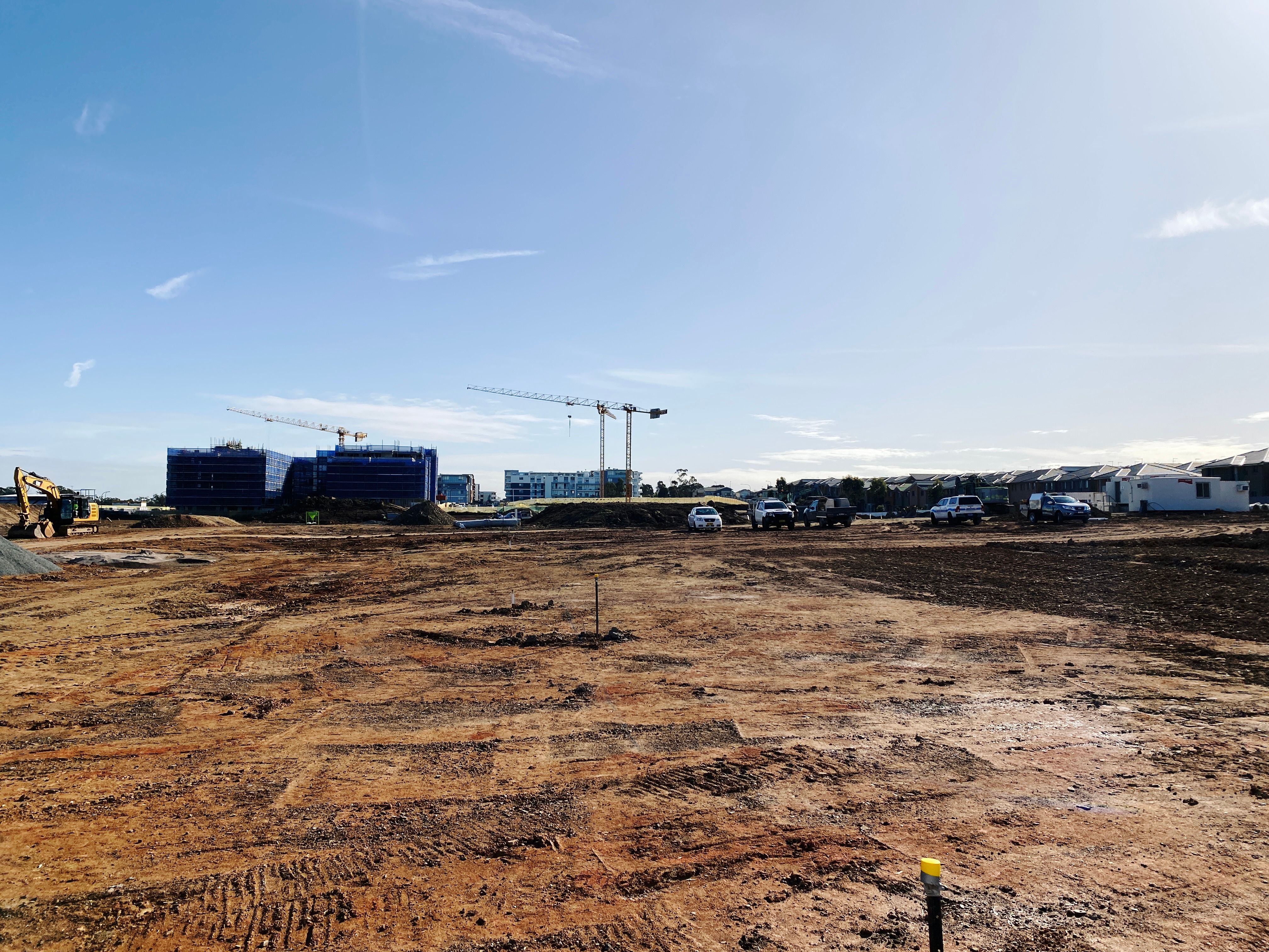 A photo of a big open field of dirt with utes parked and earthworks equipment, with big unit blocks in the distance and more partially-completely ones in front of them.