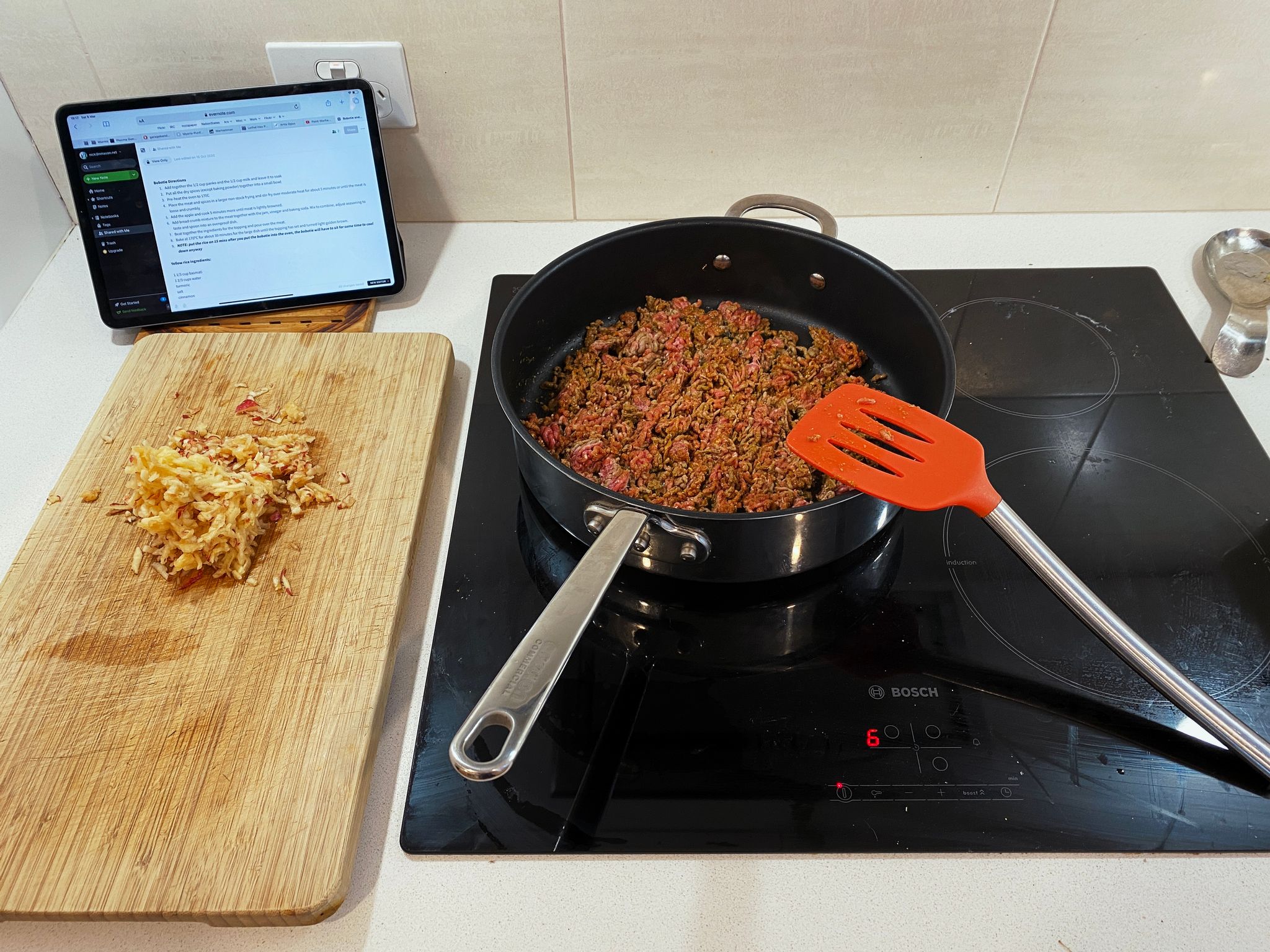 A photo of a stove with a frying pan with beef mince and lots of spices in it mid-cooking. Next to the stove is a cutting board with a grated apple on it.