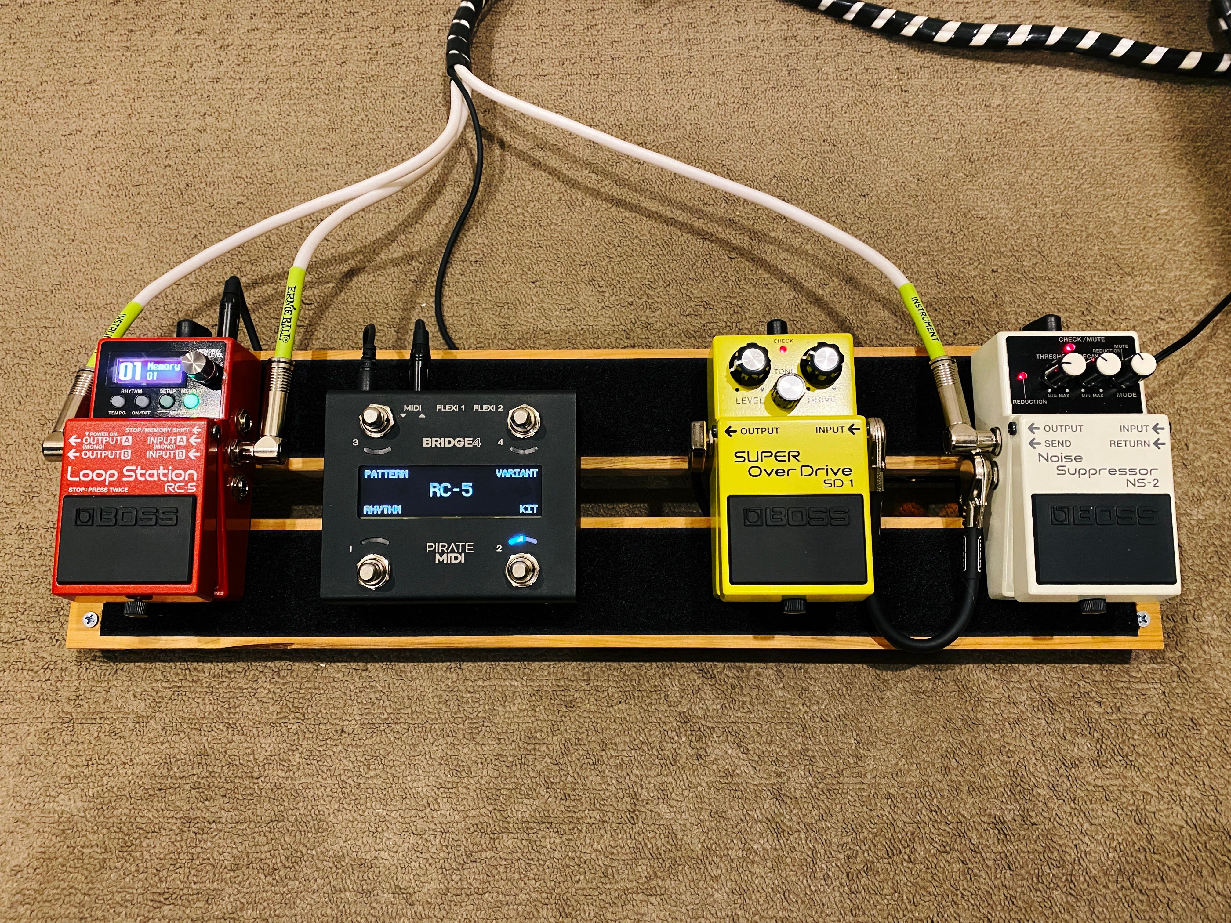 A photo of a DIY guitar pedalboard. It consists of two horizontal planks of wood screwed into two shallowly-angled pieces on either end. The two horizontal pieces have strips of velcro on them, and there's four pedals attached to it. The new pedal is black with four silver switches on it and a little LCD display in the middle.