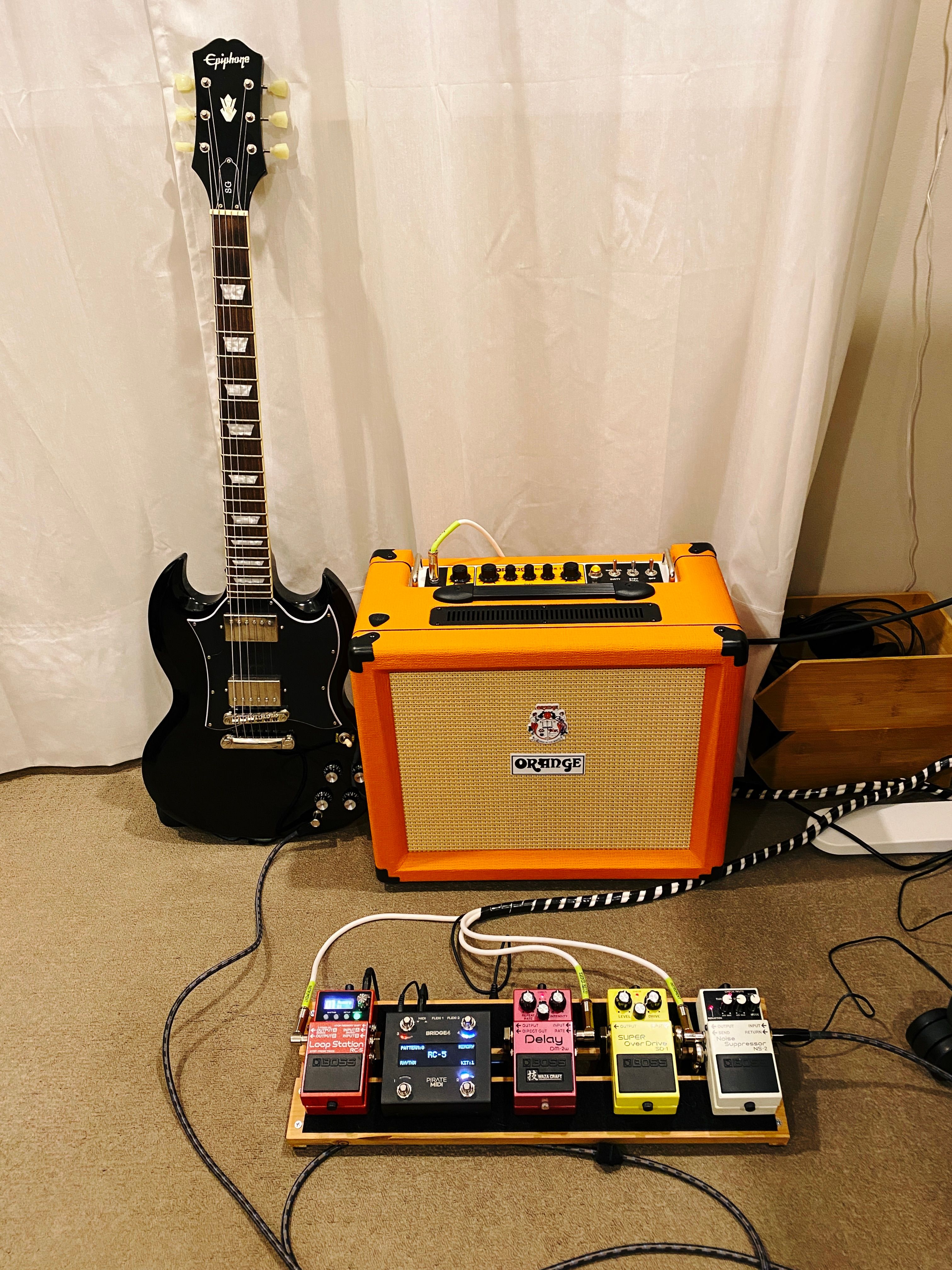 A photo of an Orange brand guitar amp (which is also actually orange-coloured, the Orange Rocker 15 Combo) sitting next to a shiny black electric guitar (an Epiphone SG Standard). In front of that on the floor is my DIY wooden guitar pedalboard with four Boss brand stomp pedals on it and a black "Pirate MIDI" brand MIDI controller pedal.