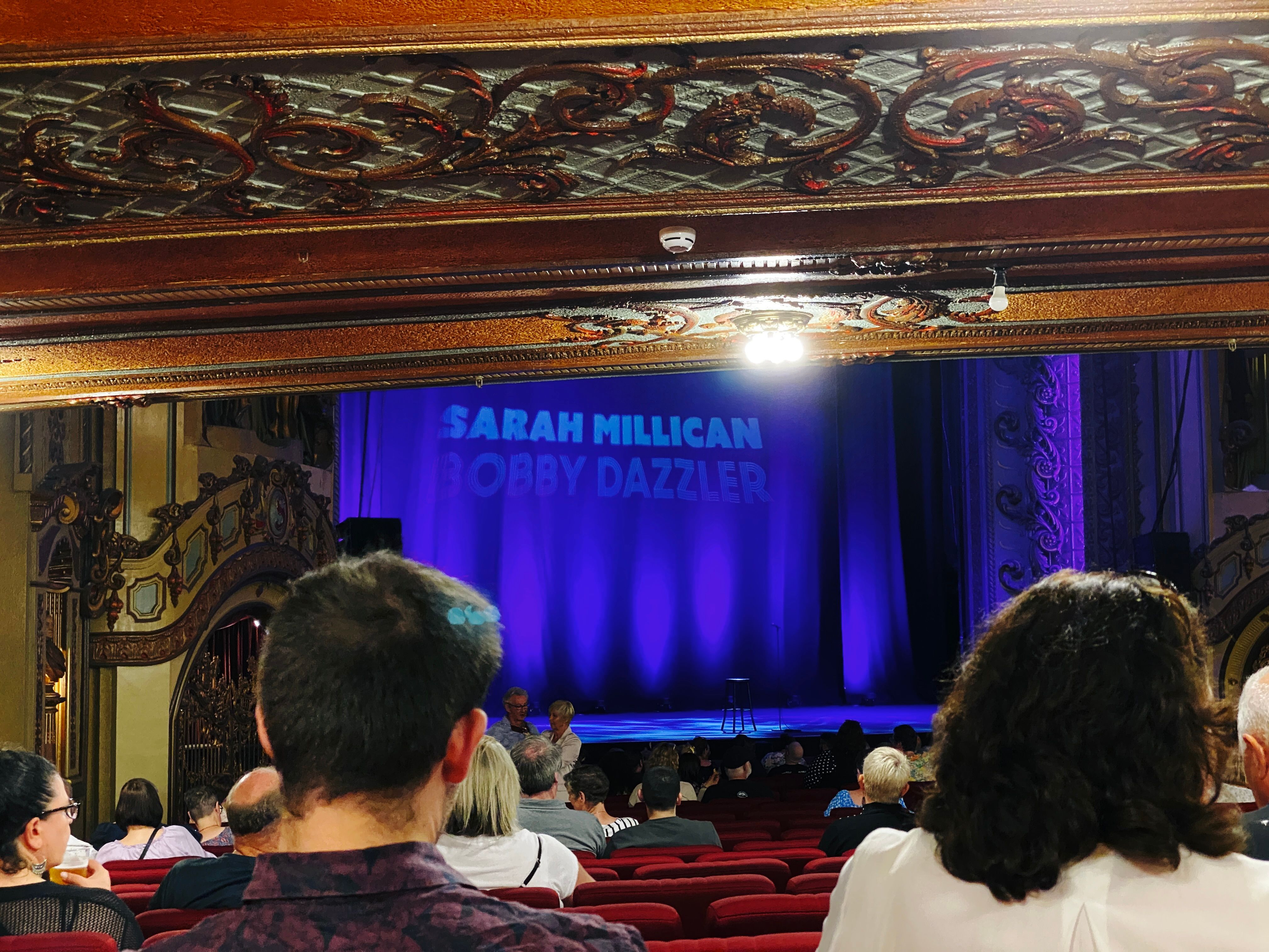 A photo looking towards the stage at Sydney's State Theatre with a big purple backdrop that says in all capitals "SARAH MILLICAN", with dimmer text underneath "BOBBY DAZZLER".