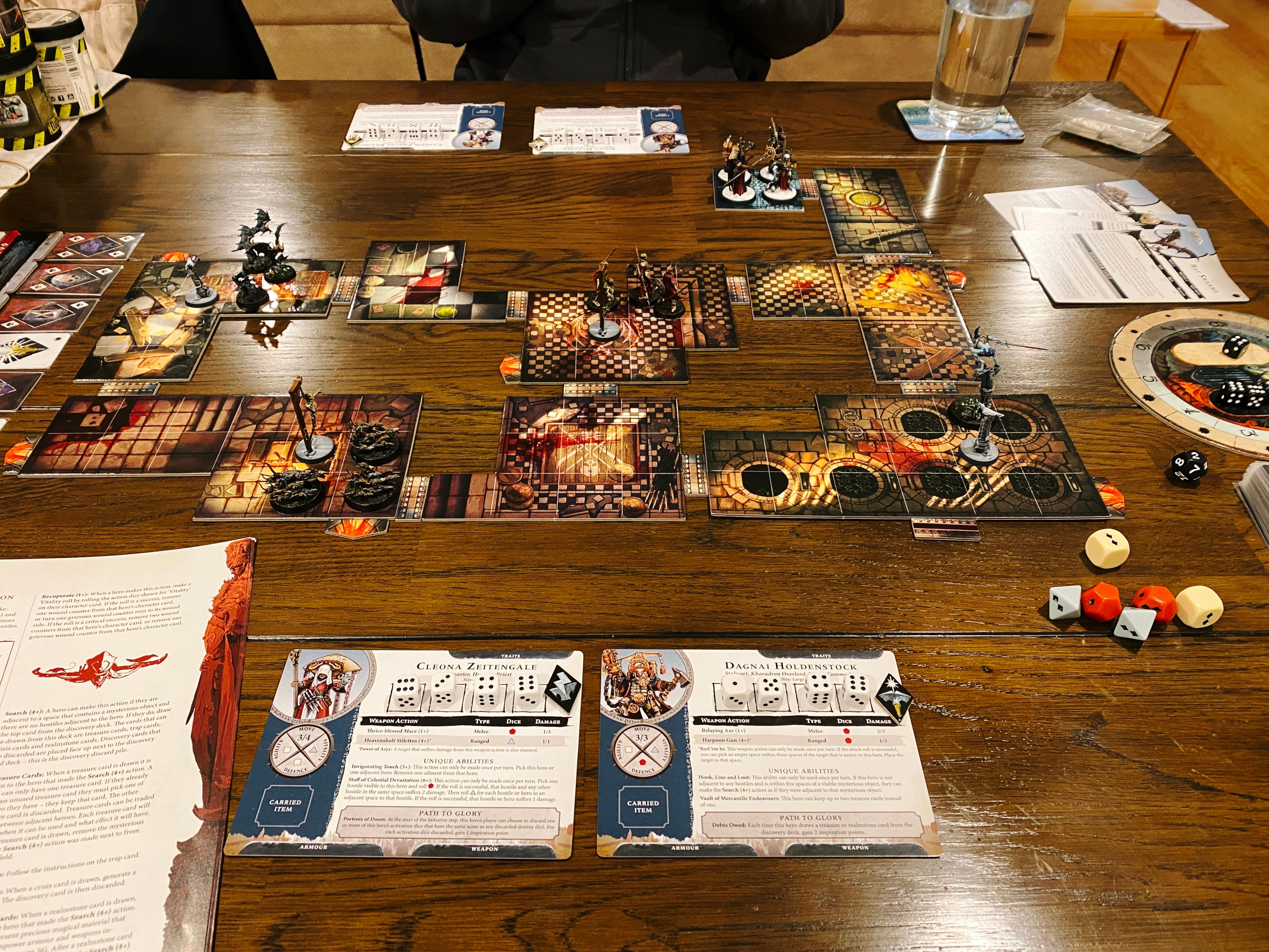 A photo of our dining table with a Warhammer Quest game set up on it. There's lots of cards and dice everywhere, and in middle are a bunch of variously-shaped tiles connected together that look like the inside of fantasy buildings. There's four hero miniatures at the top (one heavily-armoured warrior woman with a sword, a vampire hunter with a long leather coat and a big hat, a steampunk dwarf, and a priest with a rich red robe and a big staff) plus four groups of hostile miniatures at various points on the board (three skeletal warriors, three rat swarms, three bat swarms, and a lone vampire with a very long sword).