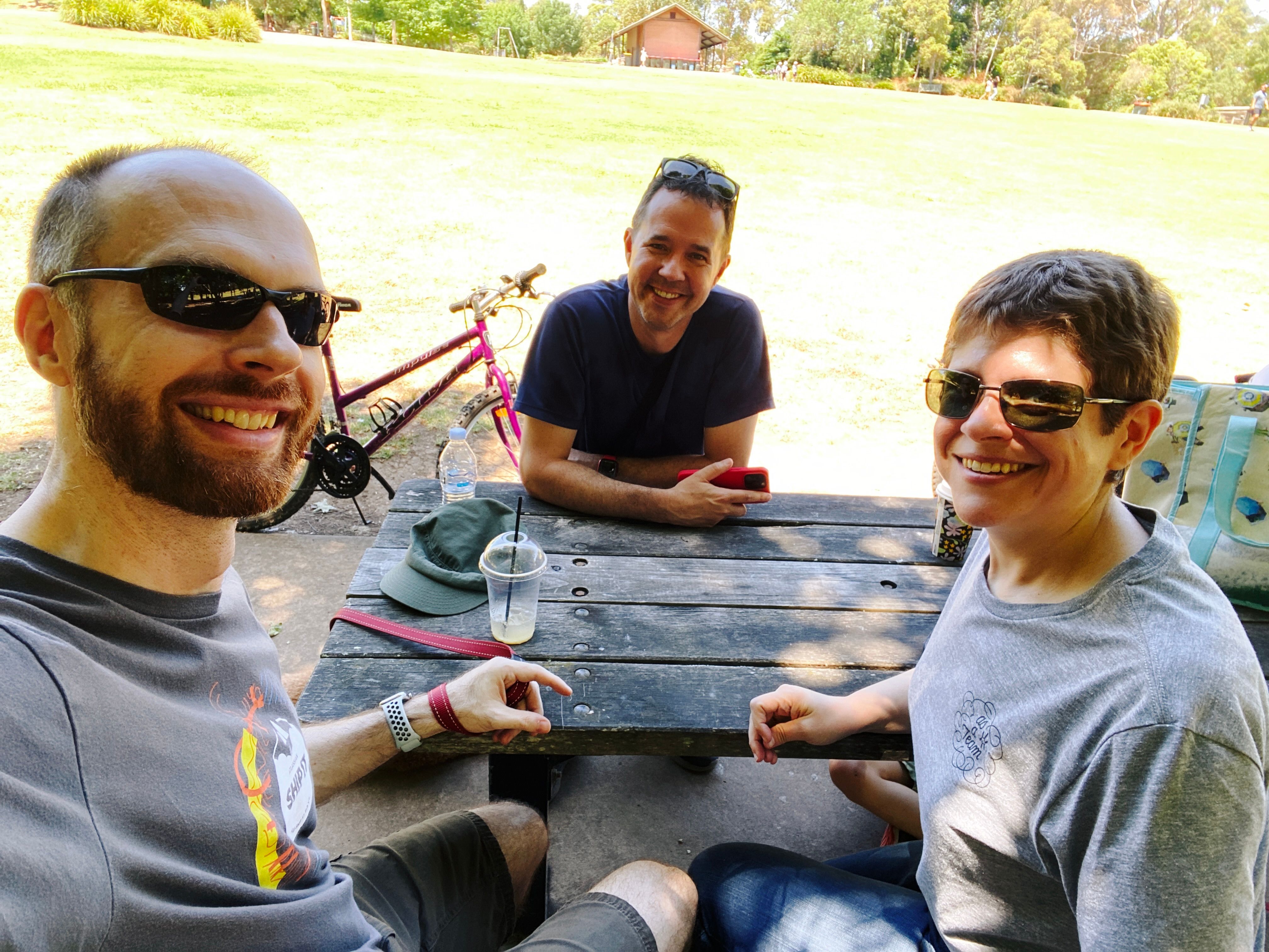 A photo of three people sitting at a park bench in the shade. There's me, a white man with a short red beard and short hair wearing sunglasses, Andrew, also a white man but without a beard, and Kristina, a white woman (also no beard) with short hair wearing sunglasses. We're all smiling at the camera.