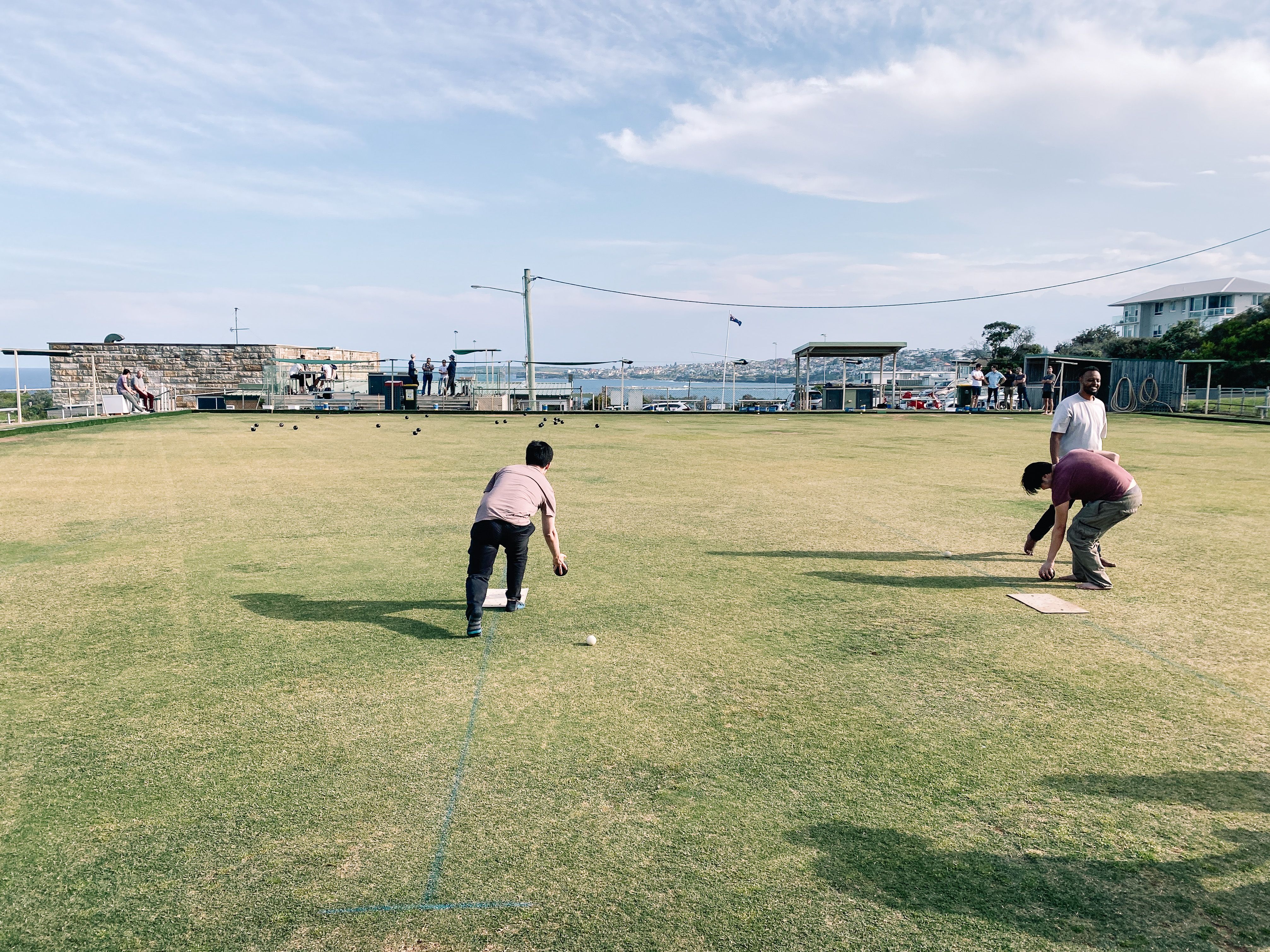 A photo looking down a lawn bowls green (very short, well-manicured grass, similar to a golf course), with a person bending down mid-bowl and a bunch of the balls already at the other end of the green close.
