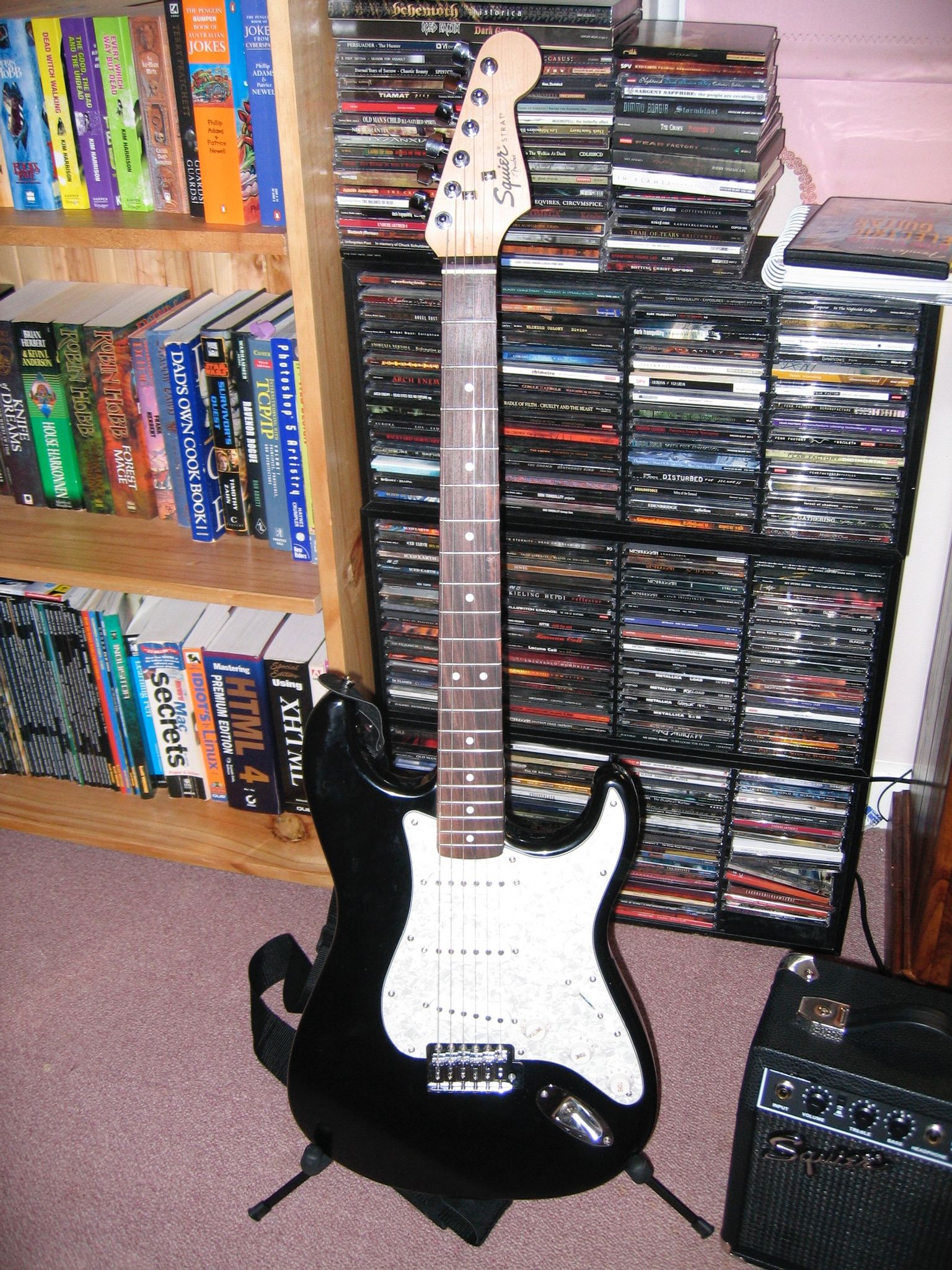 A closeup of a black electric guitar with white scratch plate sitting on a guitar stand, it has a wooden head and a strap attached.