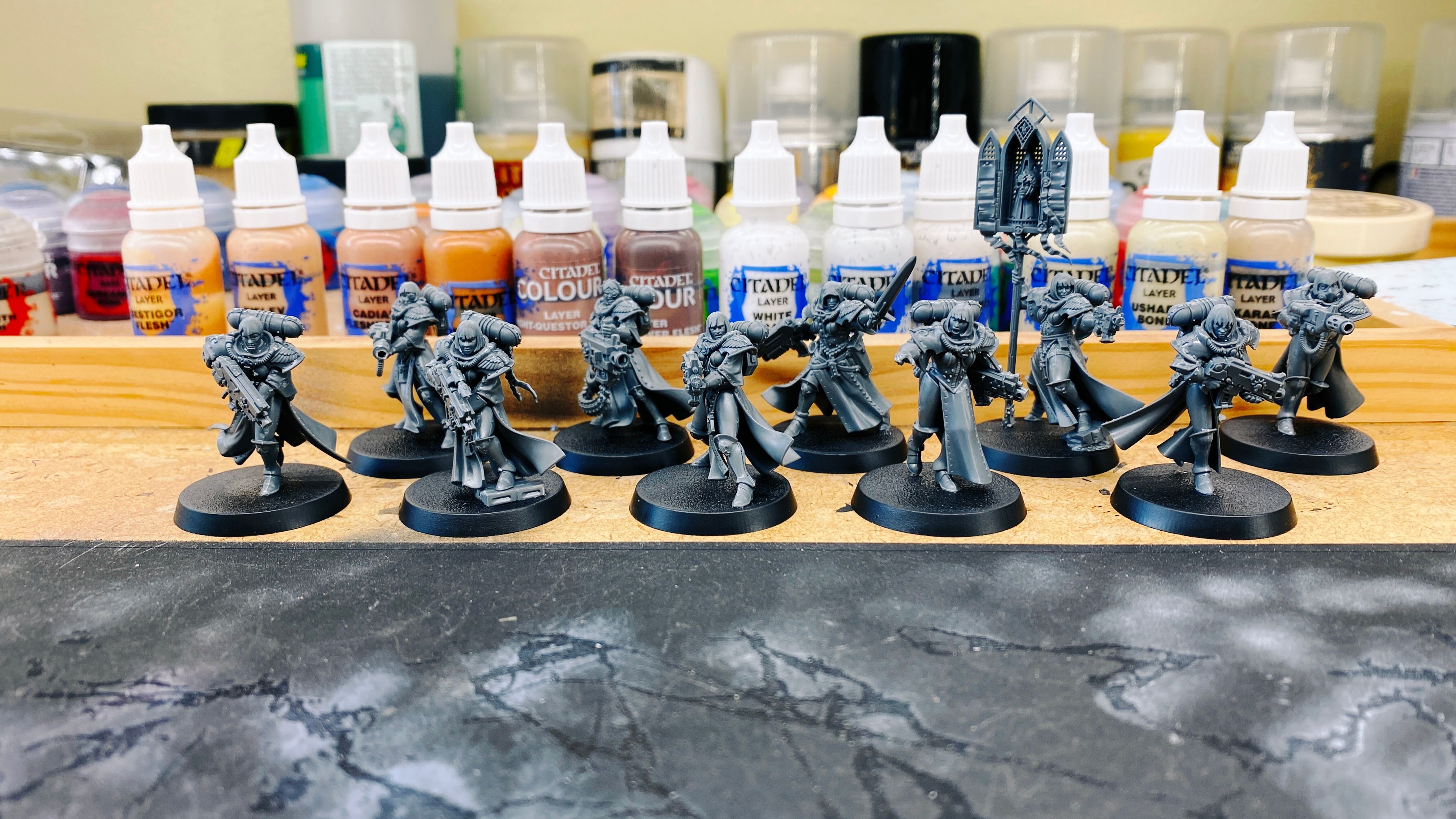 A photo of ten grey plastic miniatures of the "Sisters of Battle" faction in Warhammer 40,000. They're all women wearing power armour and wielding big-arse guns, and the lore is that they're the military wing of the Imperial Church so there's a distinctly religious feel to them.