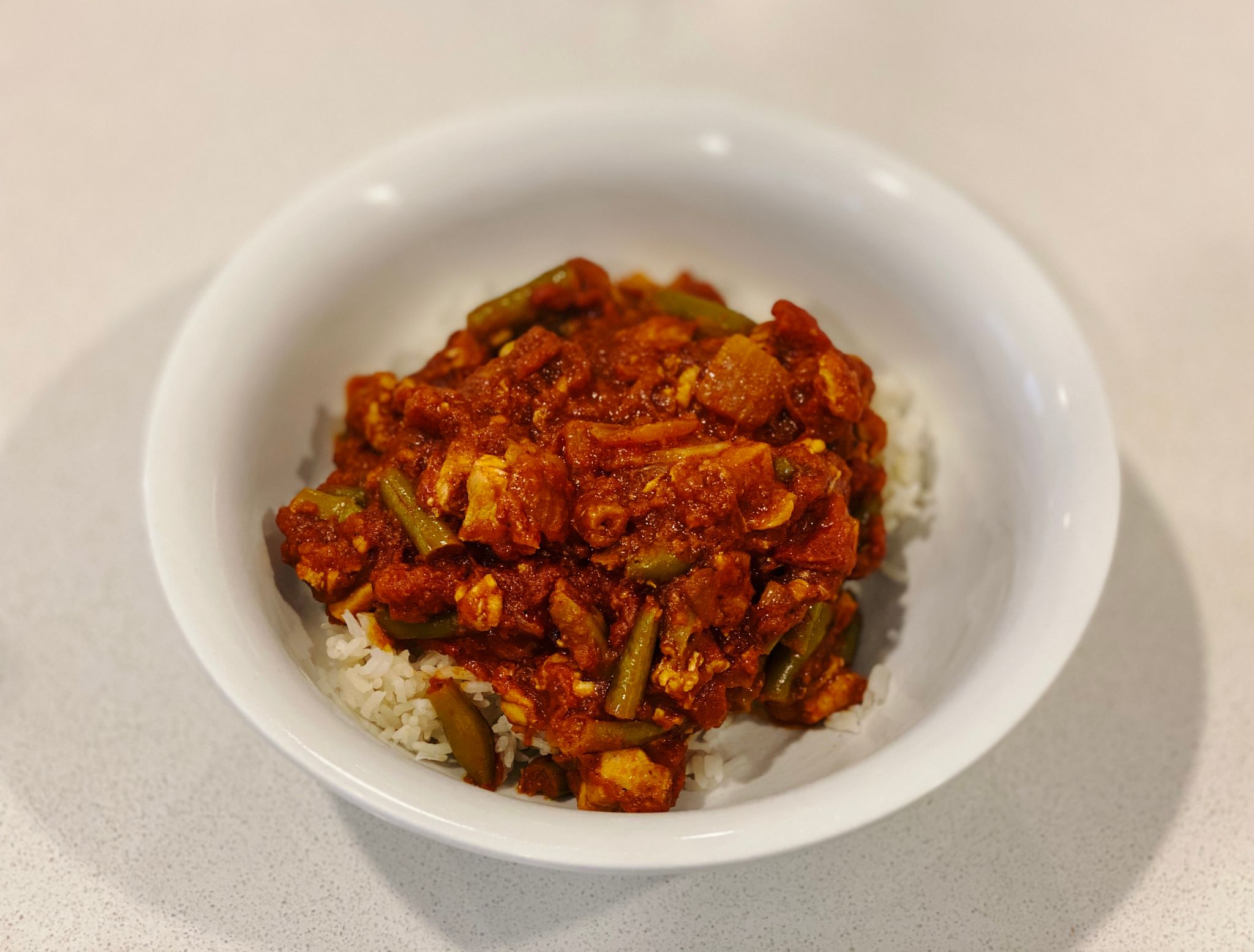 A photo of a bowl of tomato-based Indian chicken curry with green beans in it.