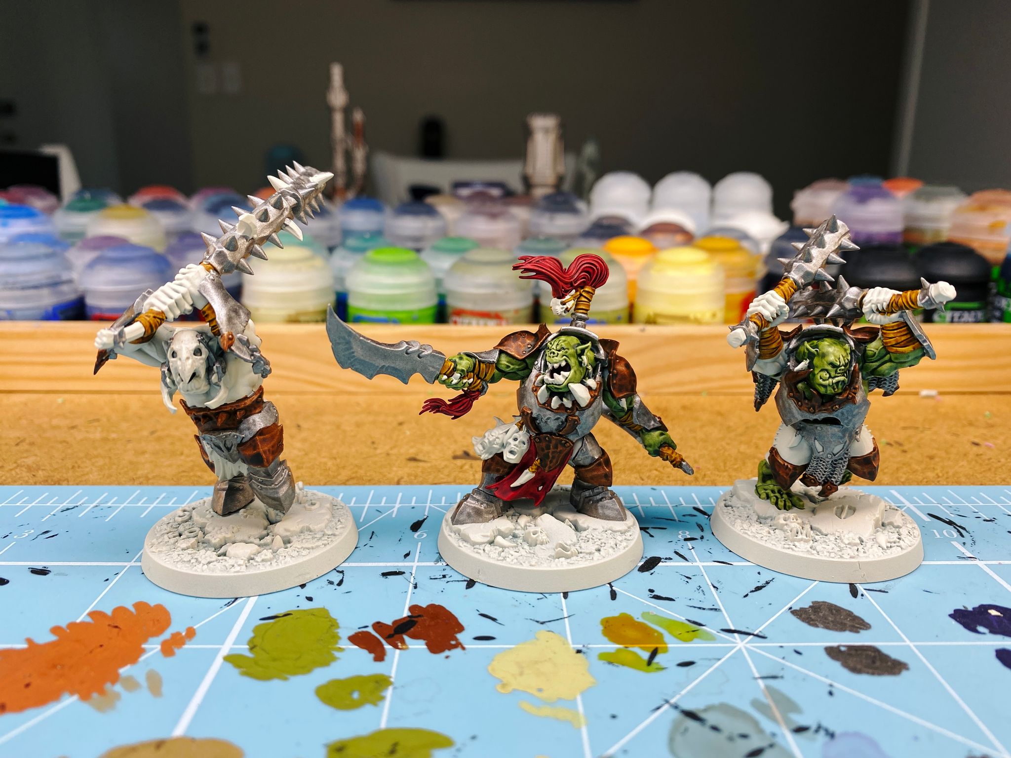 A photo of three partially painted "orruks" (Games Workshop speak for orks), the Morgok's Krushas warband from Warhammer Underworlds. They're all in battered-looking dark metal and bronze armour, the leader is pointing a sword and roaring, and the other two are mid-weapon swing. Two of the three have their green skin painted.