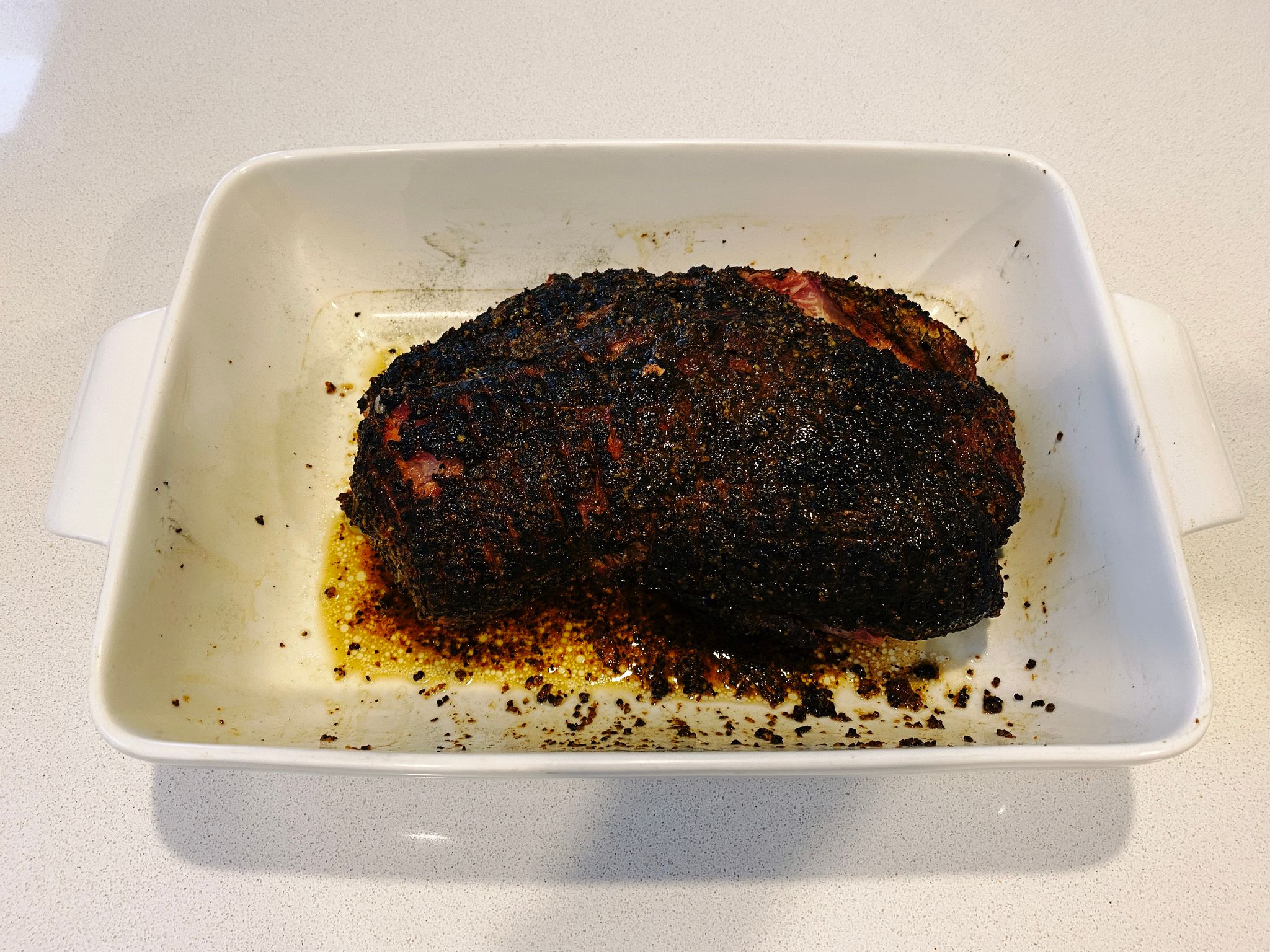 A photo of a fully cooked pork shoulder sitting in a big white dish. It's dark and has a nice crust on the outside.
