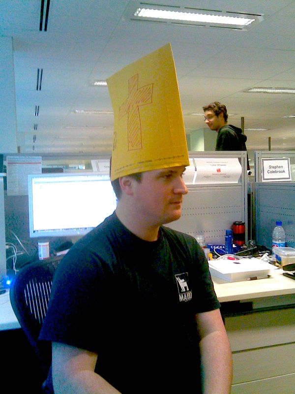 A photo of my friend Nick sitting in a chair with a yellow manila envelope opened up and sitting on his head so it looks like a hat. The envelope has a christian cross drawn on it.