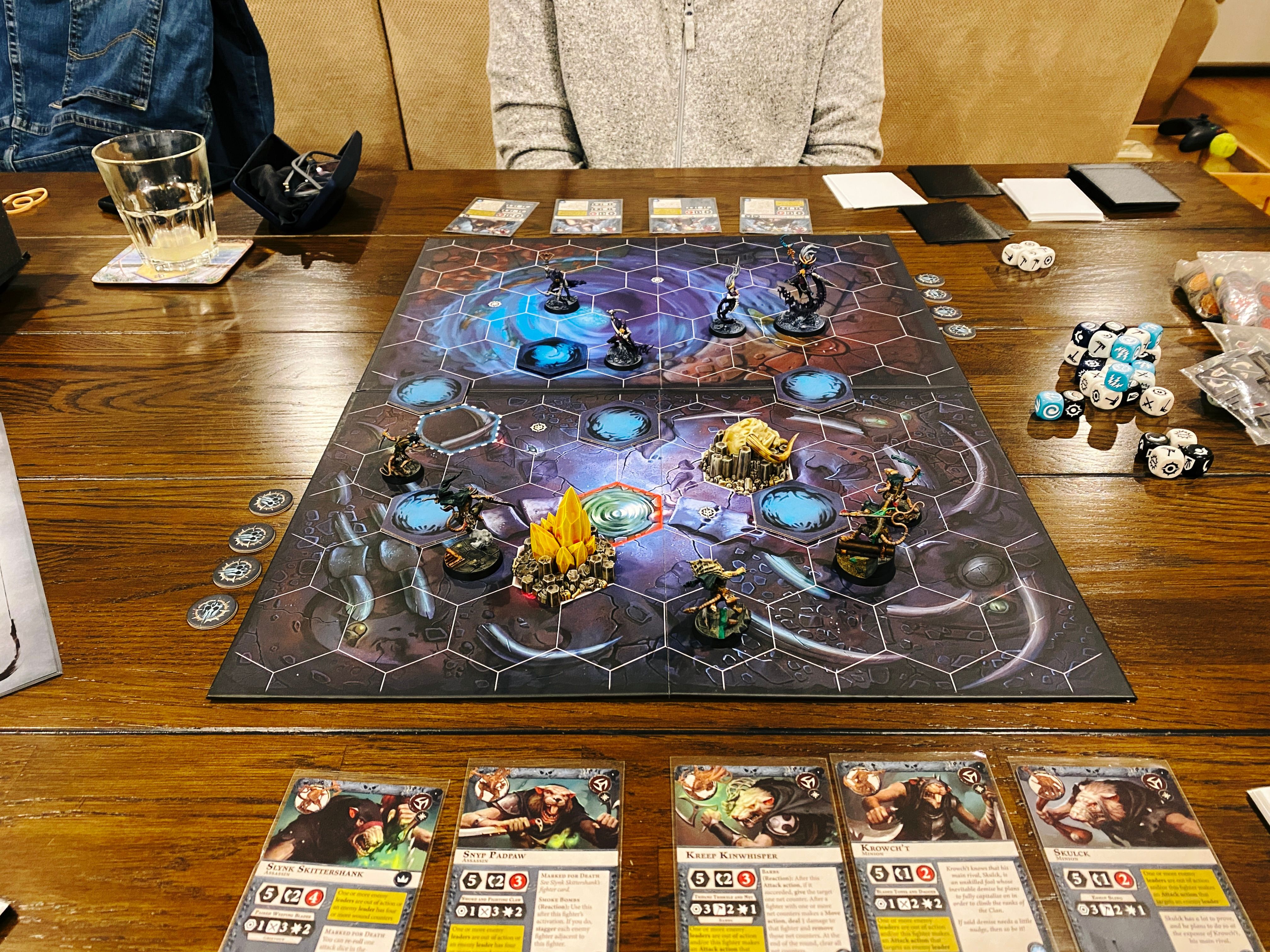 A photo of a two-player game of Warhammer Underworlds. It's played on a board with hexes on it, the artwork on the board looks like it's in a sea cave or something. Closest to the camera are five Skaven (ratmen) assassins, and facing them are four lithe elves painted mostly in deep purples.