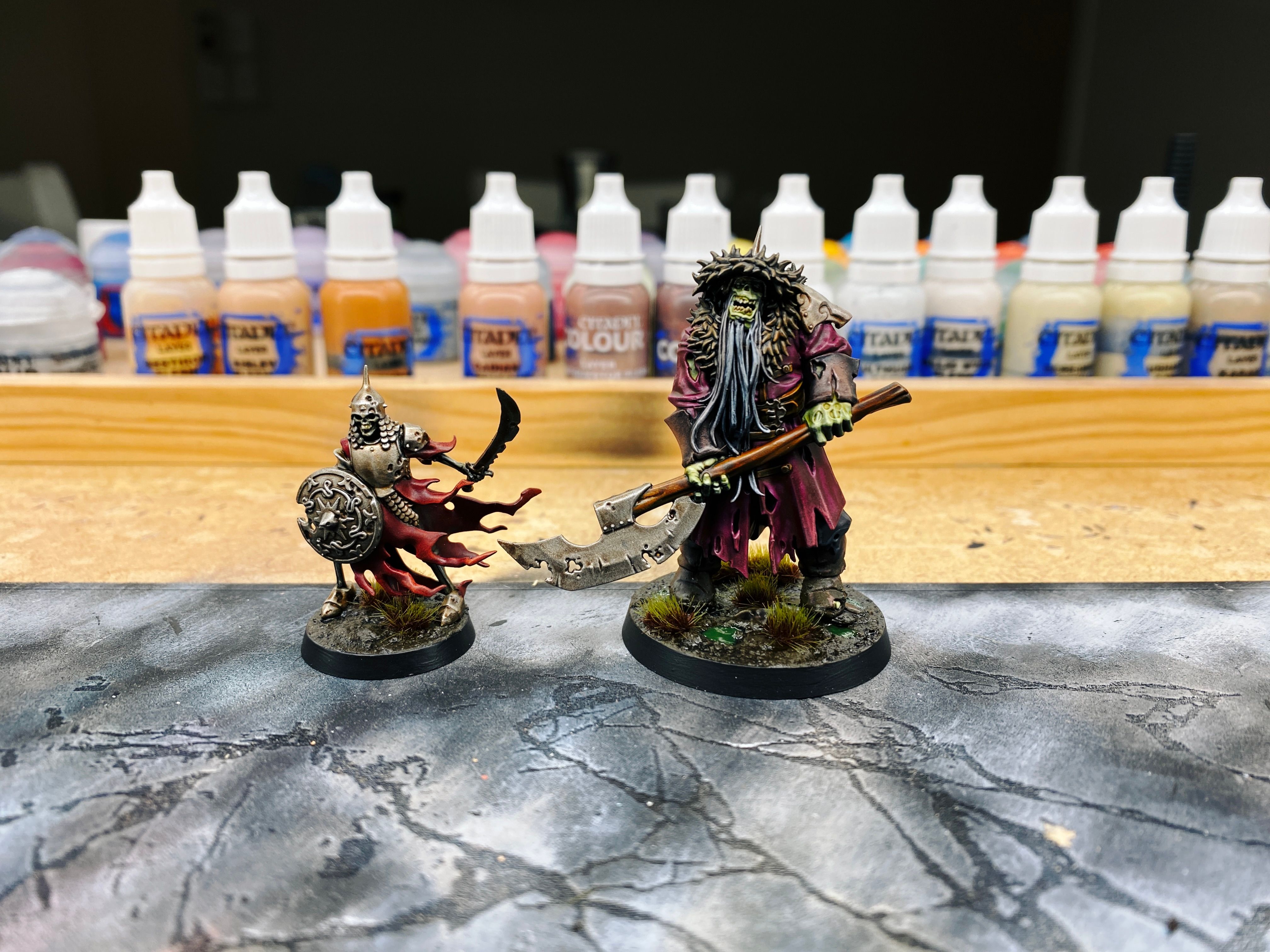 A photo of two painted miniatures, on the left is a regular human-scale skeletal warrior, but standing next to him is the positively huge Kosargi Night Guard. He's a hulking brute that's close to twice the height of the skeleton, with rotting green skin and wearing a torn-up knee-length burgundy cloak and a Mongolian-style fur hat. He's got a big long grey beard and is holding an absolutely massive axe that itself is longer than the skeleton is tall. The base looks like mud with gross green swampy water in some of the hollows, and some swampy grass tufts scattered about.