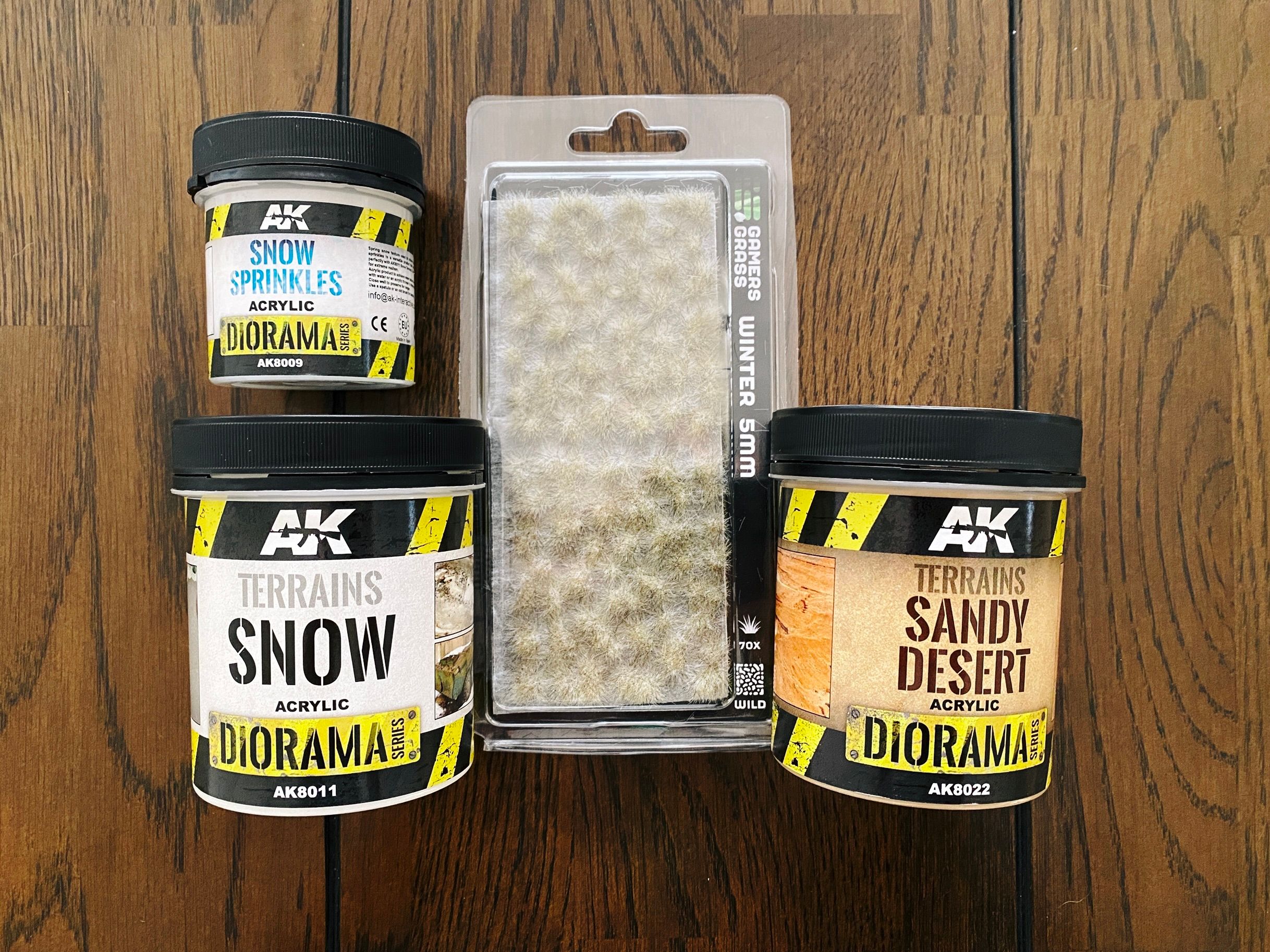 A photo of three tubs of texture paint (snow, snow sprinkles, and "Sandy Desert"), and a little packet of light green/brown grass tufts that look like they've got snow on the top of them.