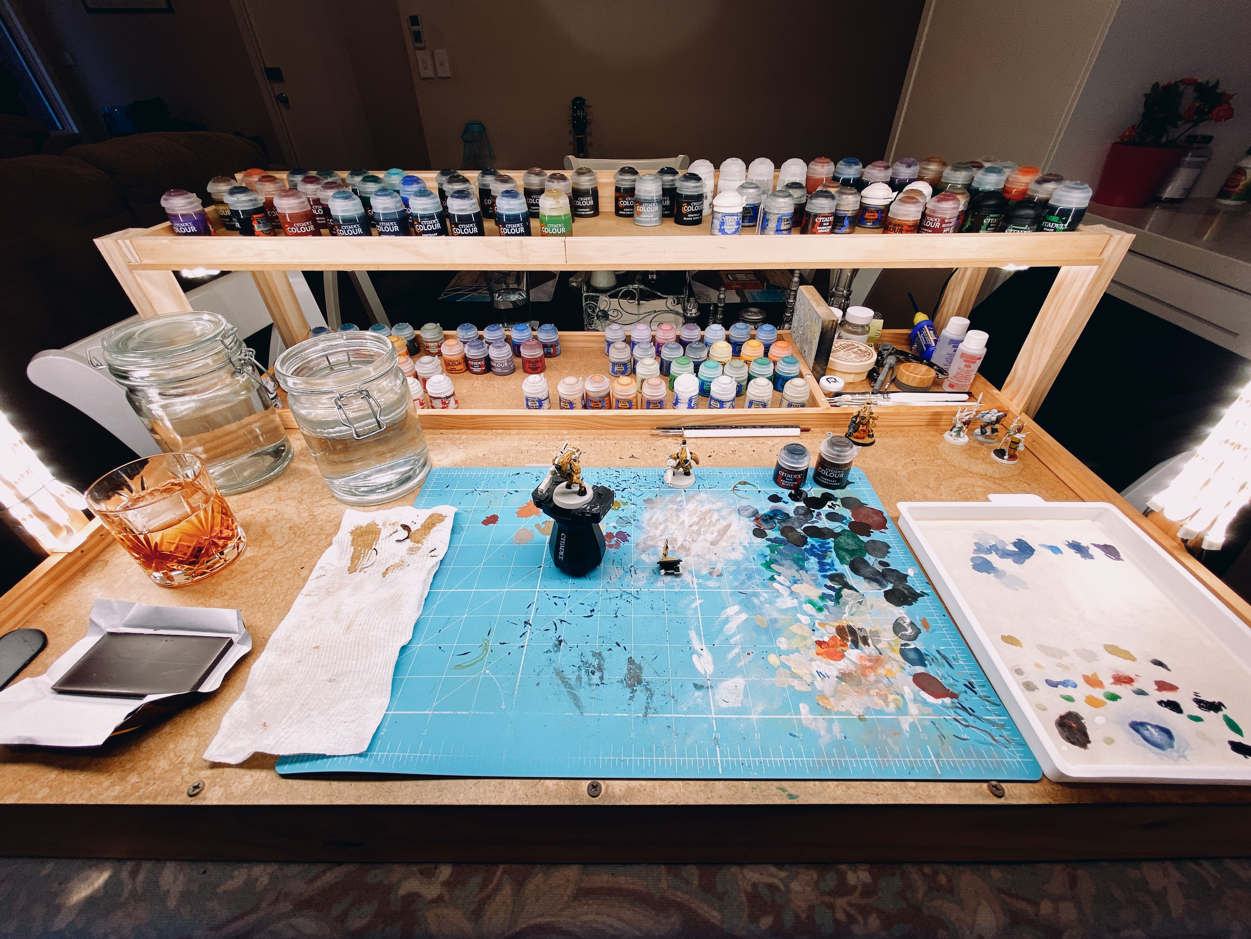 A photo of my painting table with paints all at the back, a miniature in a little holder in the middle, a wet palette at the right, and two jars of water for rinsing the brushes in at the left. There's also a glass of rum and a piece of dark chocolate at the left.